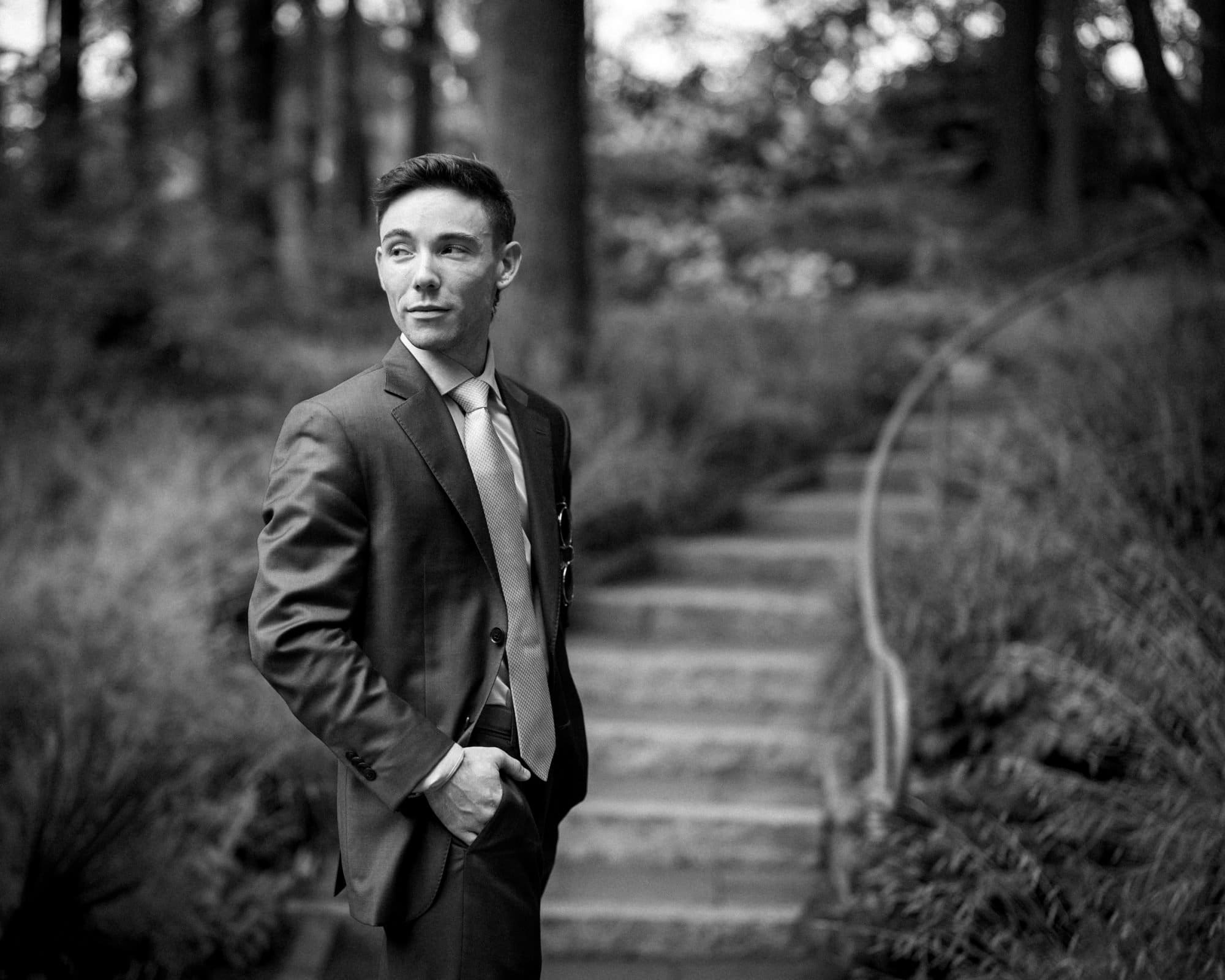 duke university wedding photography - sarah & case A man in a suit stands outdoors with one hand in his pocket, gazing to the side. Behind him, stone steps and lush greenery create a serene backdrop reminiscent of Duke University's elegant charm. The image, captured in timeless black and white, evokes the sophistication of wedding photography.