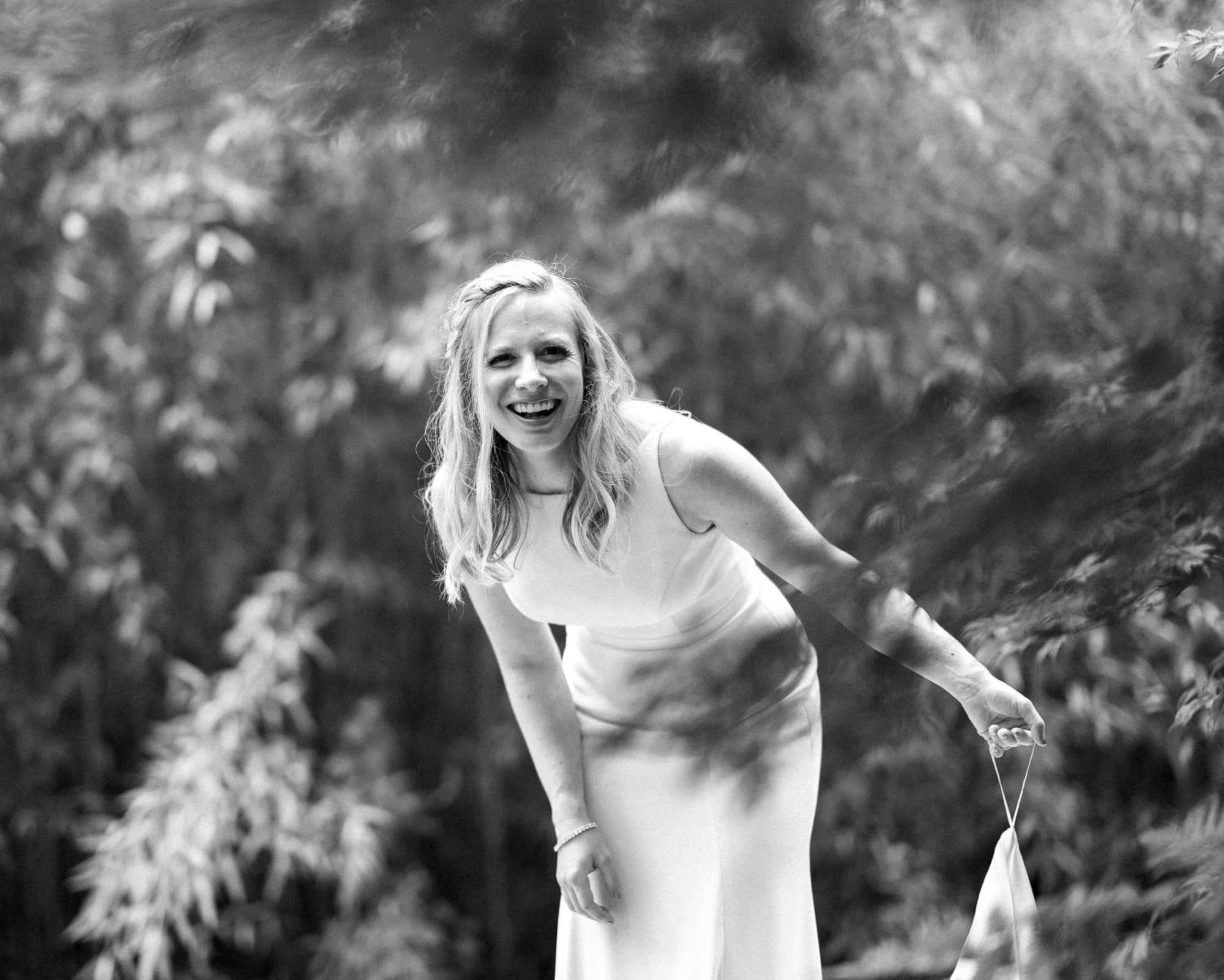 duke university wedding photography - sarah & case Black and white image of a woman in a sleeveless dress, smiling and leaning forward in a lush garden setting reminiscent of Duke University's serene landscapes. She holds a small bag in one hand, evoking the grace and elegance often captured in wedding photography.