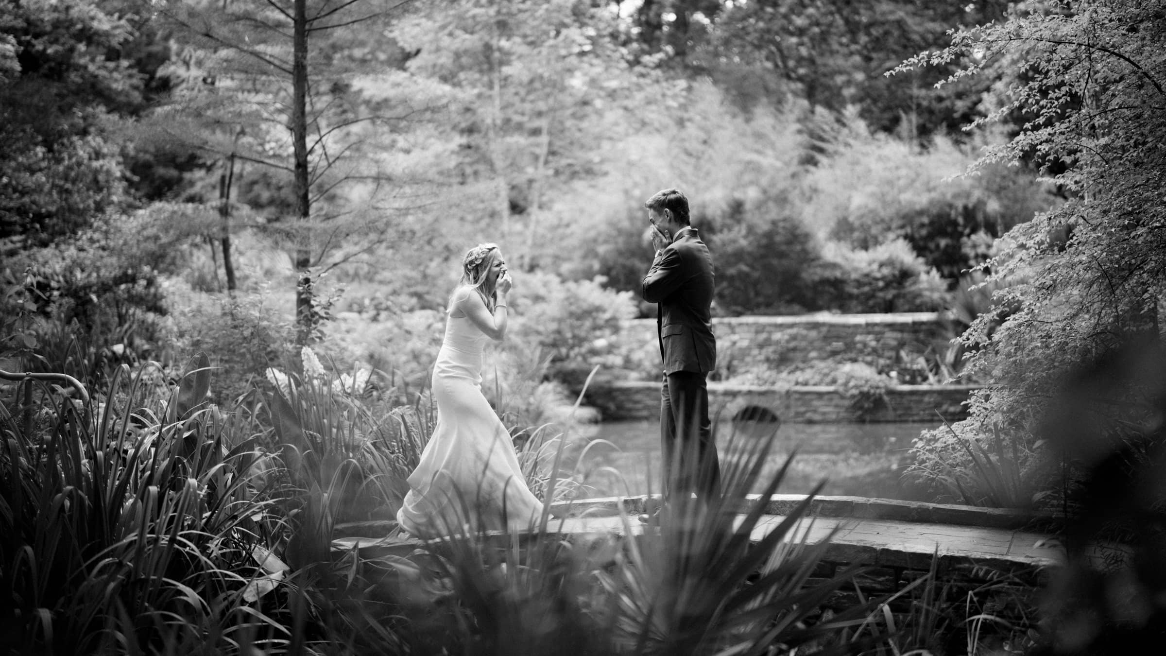 A bride and groom stand facing each other on a narrow wooden bridge enveloped by lush greenery. The bride looks joyful and surprised, while the groom stands with arms crossed, smiling. This serene, intimate scene is beautifully captured in black and white, highlighting the artistry of Duke University wedding photography.