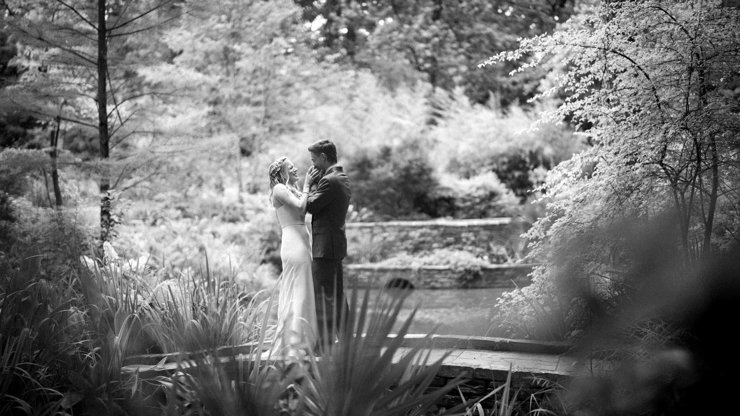 duke university wedding photography - sarah & case A black and white photo captures a couple embracing on a wooden bridge at Duke University's lush garden. The bride, in a flowing gown, and the groom, in a suit, are surrounded by dense foliage and tall trees, creating an enchanting scene of wedding photography.