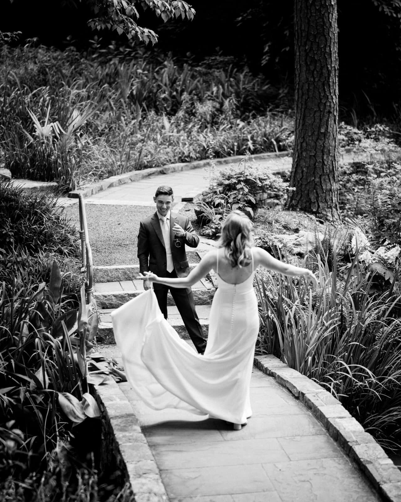 duke university wedding photography - sarah & case A black-and-white photo captures a bride and groom at Duke University. The bride, in a flowing gown, joyfully twirls on a stone path lined with plants as the groom, in his suit, extends his hand. Surrounded by greenery and trees, this moment of wedding bliss is timelessly preserved.