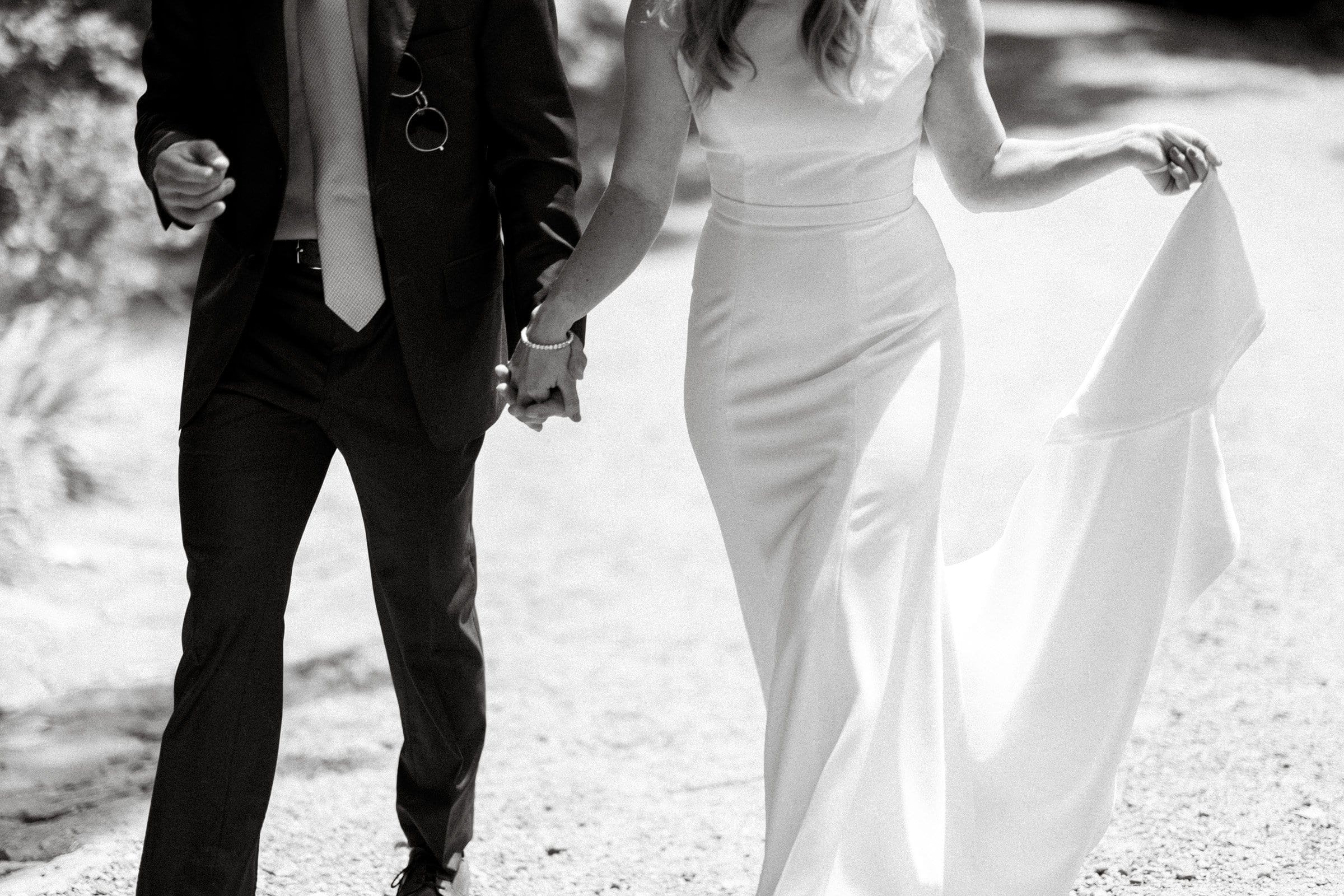 duke university wedding photography - sarah & case A bride and groom walk hand in hand down a sunlit path at Duke University. The bride wears a flowing white dress, and the groom is in a dark suit with a light tie. Their faces are not visible, capturing a candid moment of joy and connection through expert wedding photography.