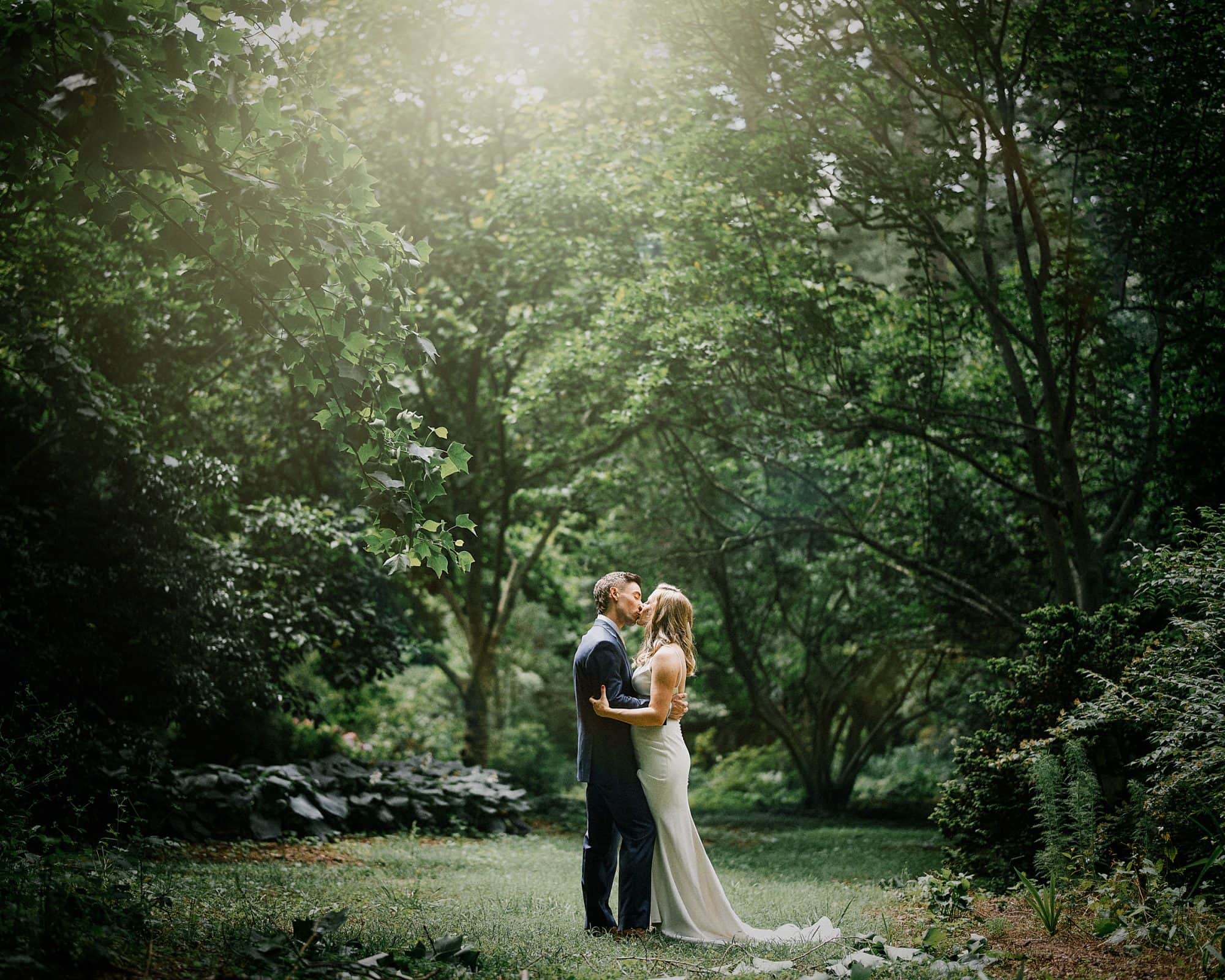 duke university wedding photography - sarah & case A couple embraces in a lush garden at Duke University, under dappled sunlight. The man, in a suit, gently kisses the woman, who wears a long white dress. Trees and bushes surround them, creating a serene and romantic atmosphere perfect for wedding photography.