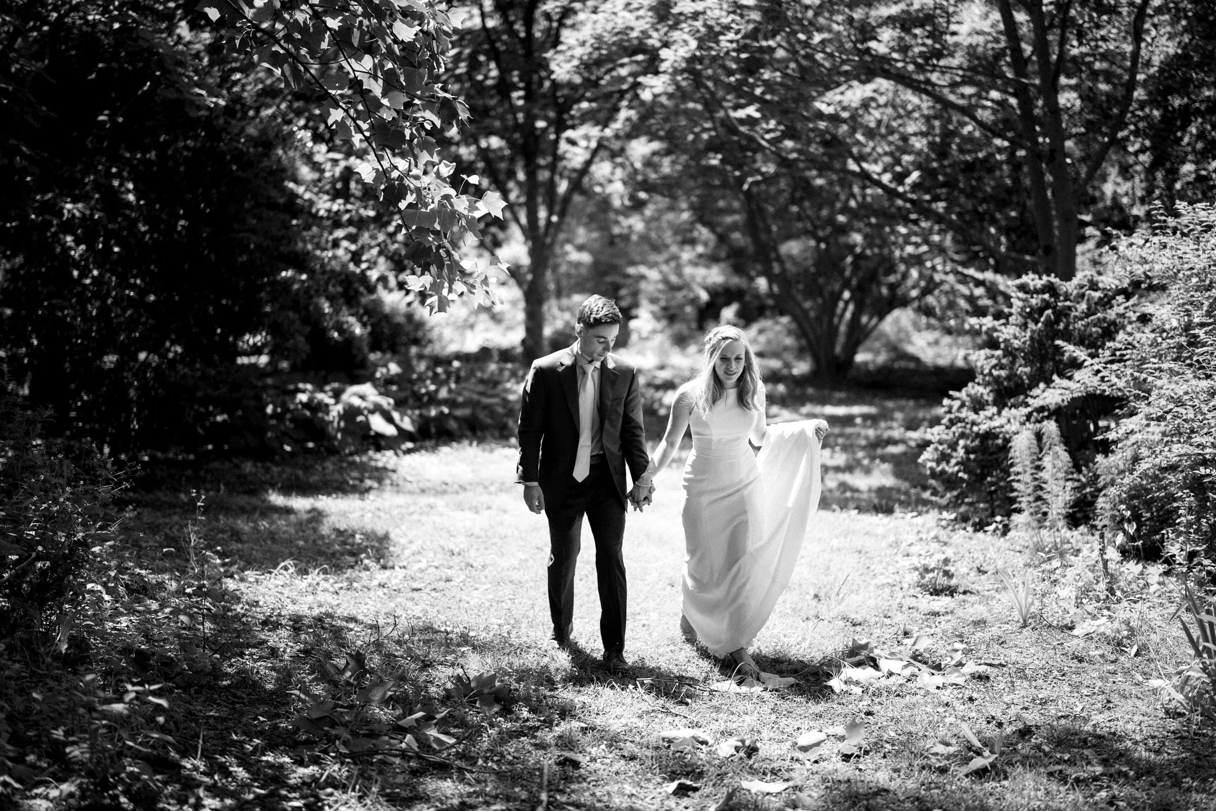 duke university wedding photography - sarah & case A black and white photo captures the essence of Duke University wedding photography, with a couple walking hand in hand through a sunlit, grassy area surrounded by trees. The woman in a long dress and the man in a suit appear relaxed and happy amidst the serene, natural setting.