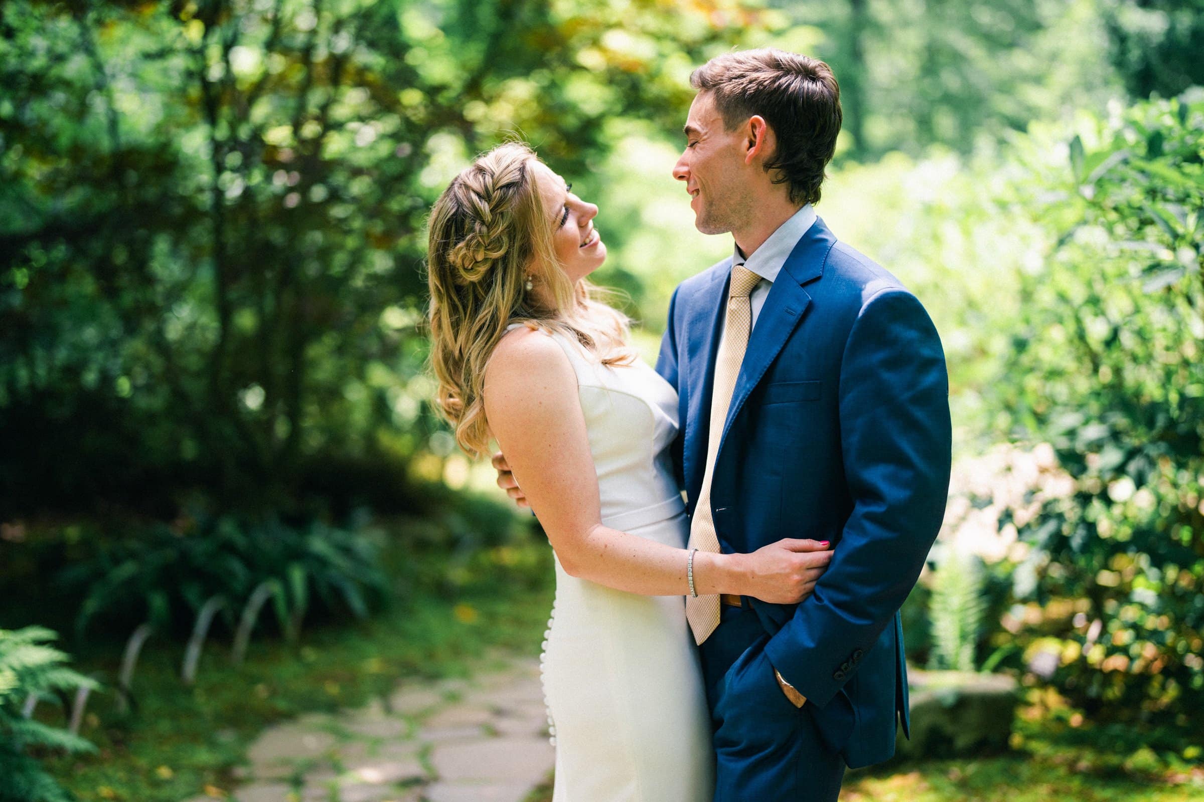 duke university wedding photography - sarah & case A couple embraces on a stone path in a lush garden, captured perfectly in Duke University's renowned wedding photography style. The woman wears a white dress, and the man dons a blue suit with a beige tie. They gaze into each other's eyes, surrounded by greenery and dappled sunlight.