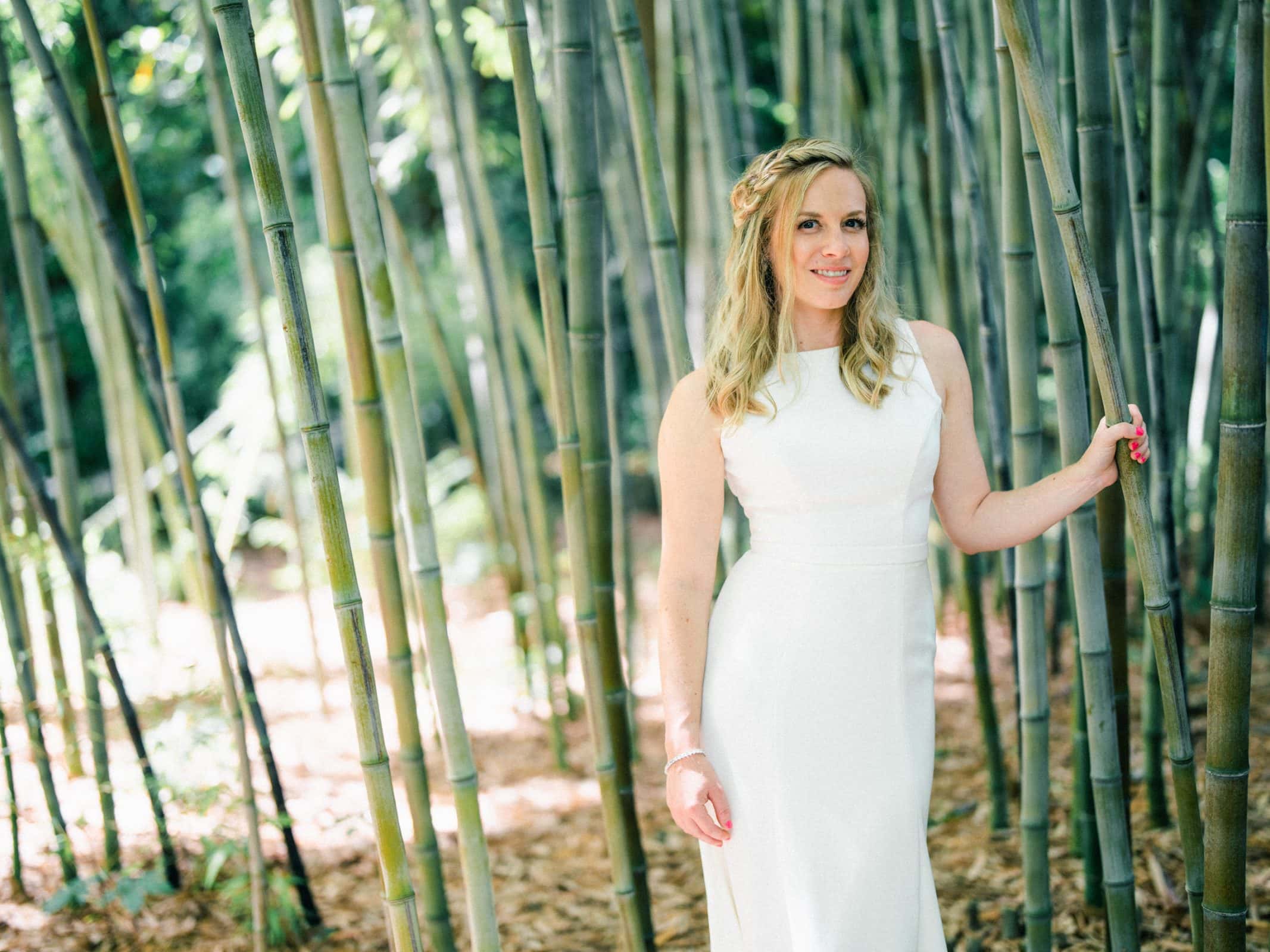 duke university wedding photography - sarah & case A woman in a white dress stands among tall bamboo stalks in a sunlit forest, her light hair glowing as she smiles with one hand resting on a bamboo stalk. The scene captures the elegance of Duke University's renowned wedding photography style, with the ground covered in dry leaves.