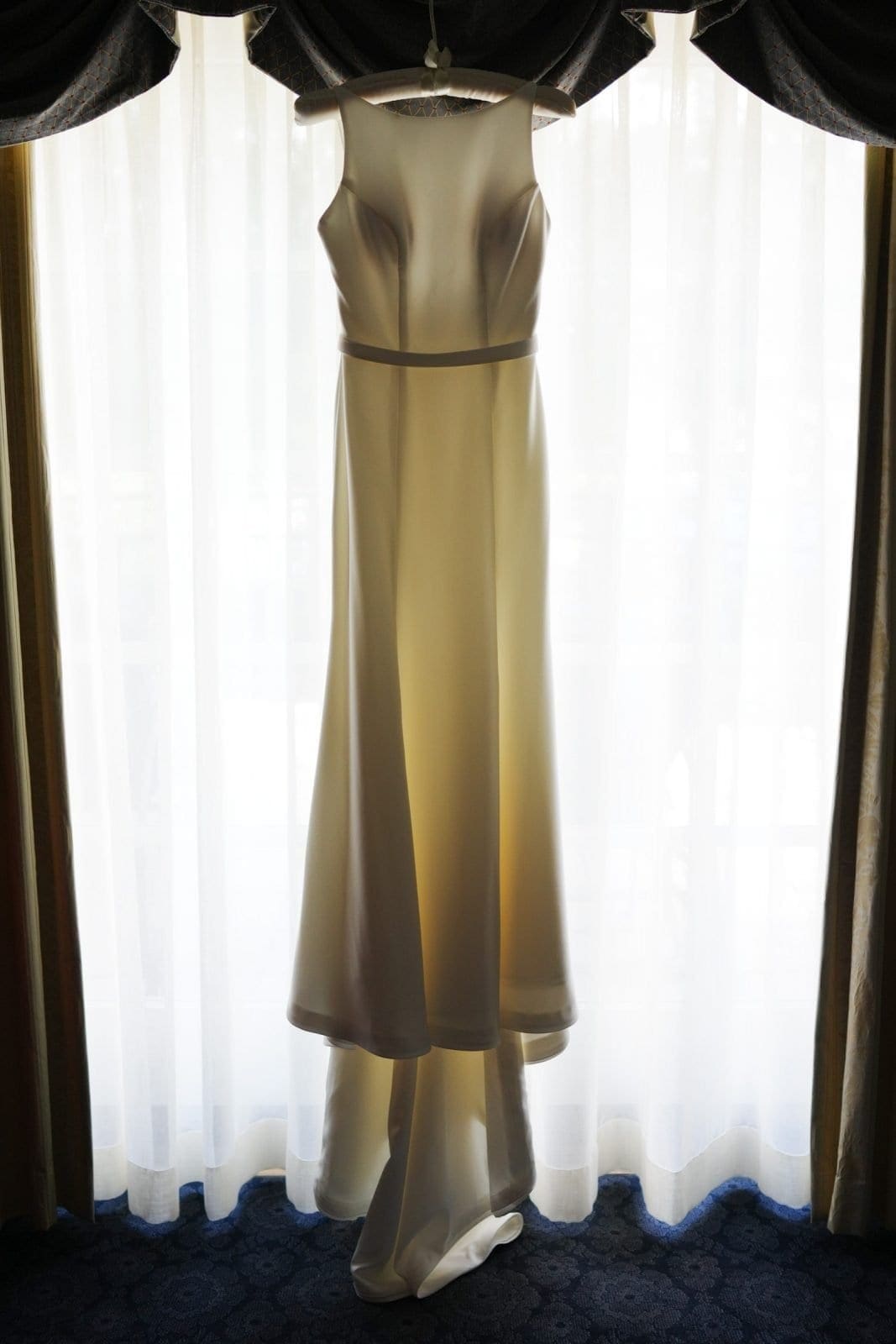 duke university wedding photography - sarah & case A white wedding dress with a simple design is hanging by a window, illuminated by natural light. Captured in the style of Duke University wedding photography, the floor-length gown features a fitted bodice and a flowing skirt, framed by dark curtains.