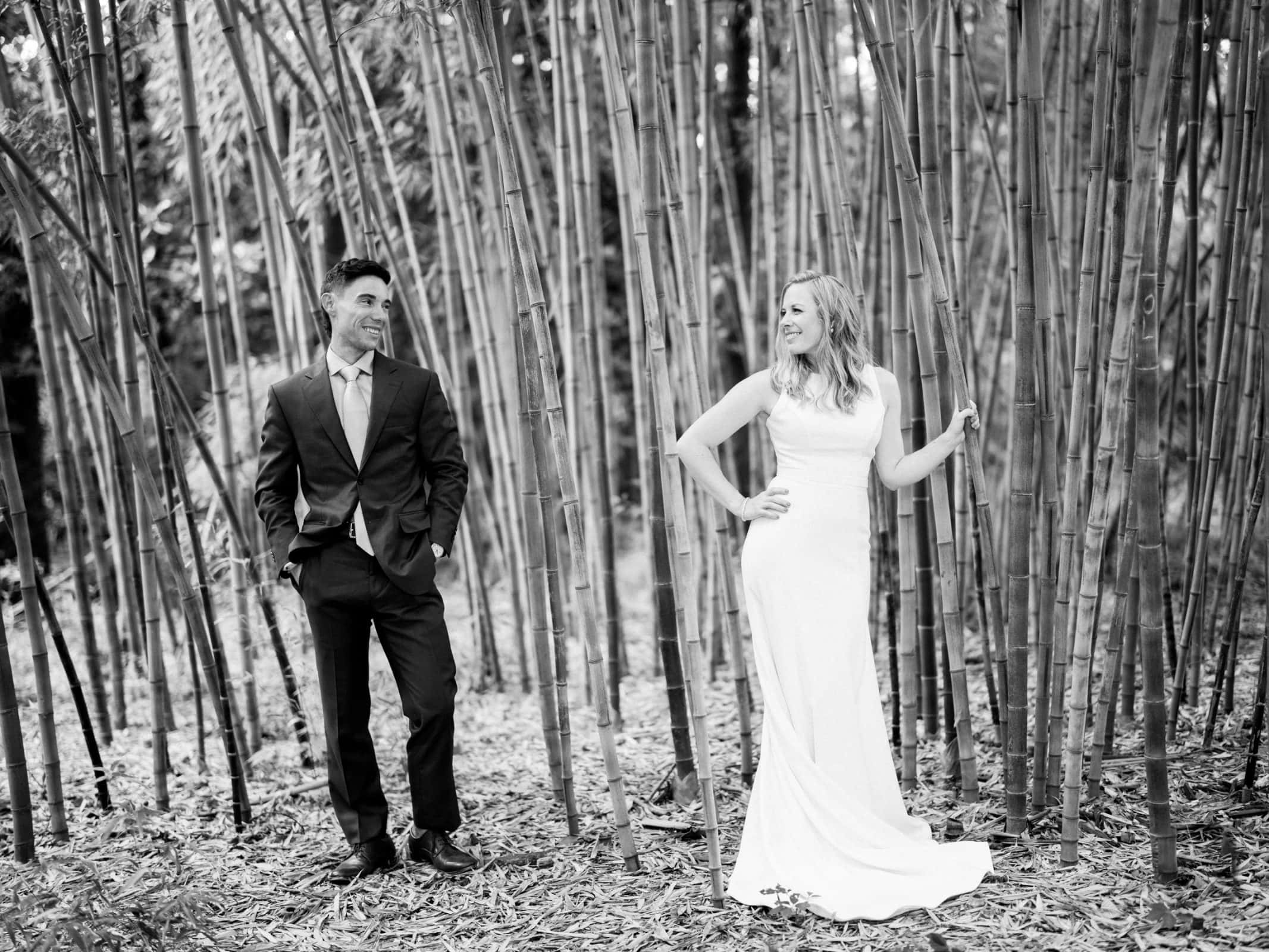 duke university wedding photography - sarah & case A man and woman stand among the tall bamboo stalks, embodying a timeless Duke University wedding photography scene. The man, in a suit, stands with hands in pockets, while the woman, in a long white dress, smiles at him with one hand on her hip and the other gently touching the bamboo. The scene is captured in black and white.