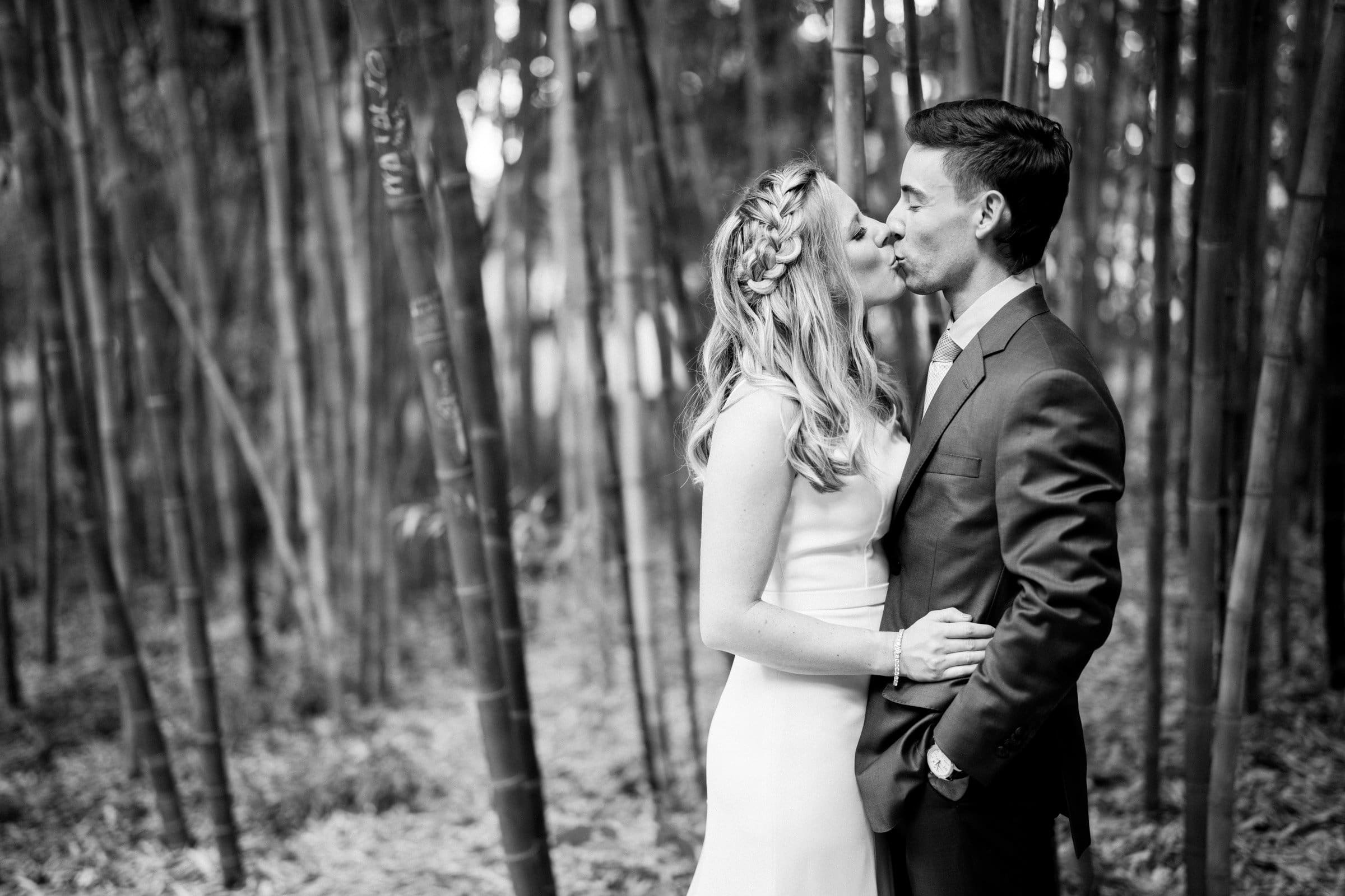 duke university wedding photography - sarah & case A black and white photo captures a couple in formal attire standing close together in a bamboo grove. The woman, in a white dress with braided hair, gazes at the man in a suit, who is looking back at her. This serene and intimate moment evokes the elegance of Duke University wedding photography.