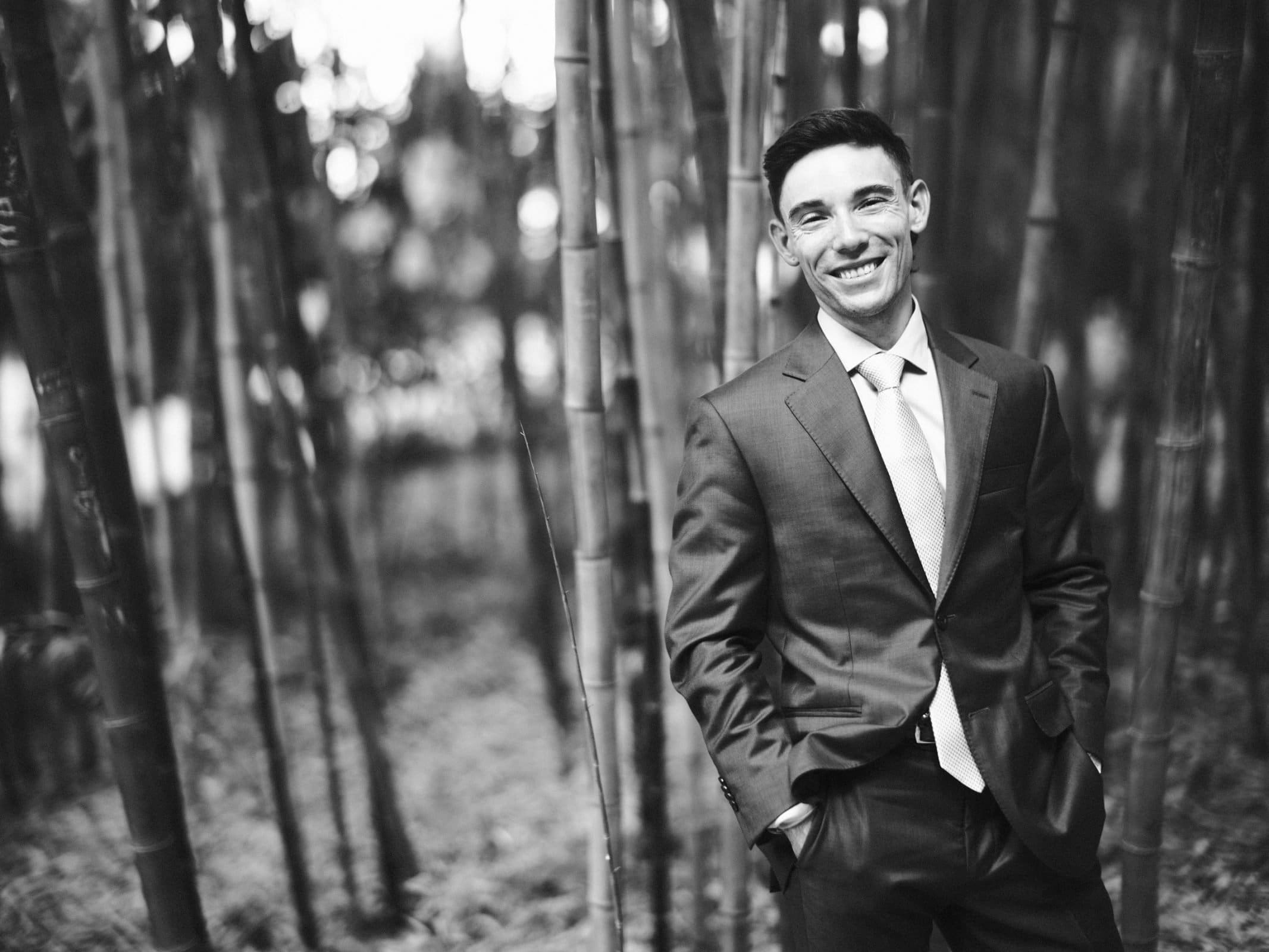 duke university wedding photography - sarah & case A man in a suit stands smiling among tall bamboo stalks in a forest setting, reminiscent of classic Duke University wedding photography. The black-and-white image highlights the striking contrast between his formal attire and the natural surroundings.