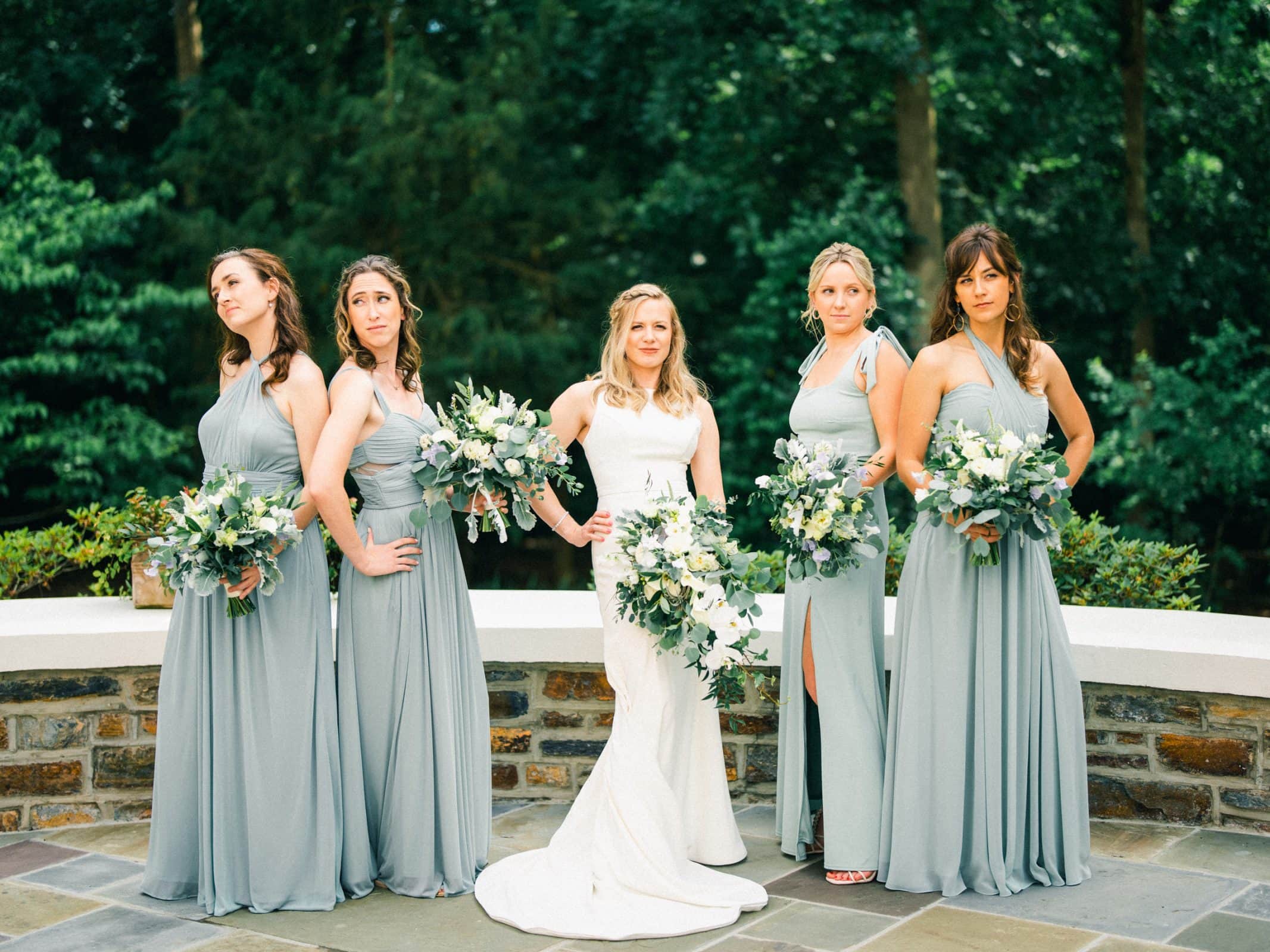 duke university wedding photography - sarah & case A bride in a white dress stands with four bridesmaids in light gray dresses, each holding a bouquet of white and green flowers. They pose outdoors against a lush, green forest backdrop—an enchanting Duke University wedding photography moment.