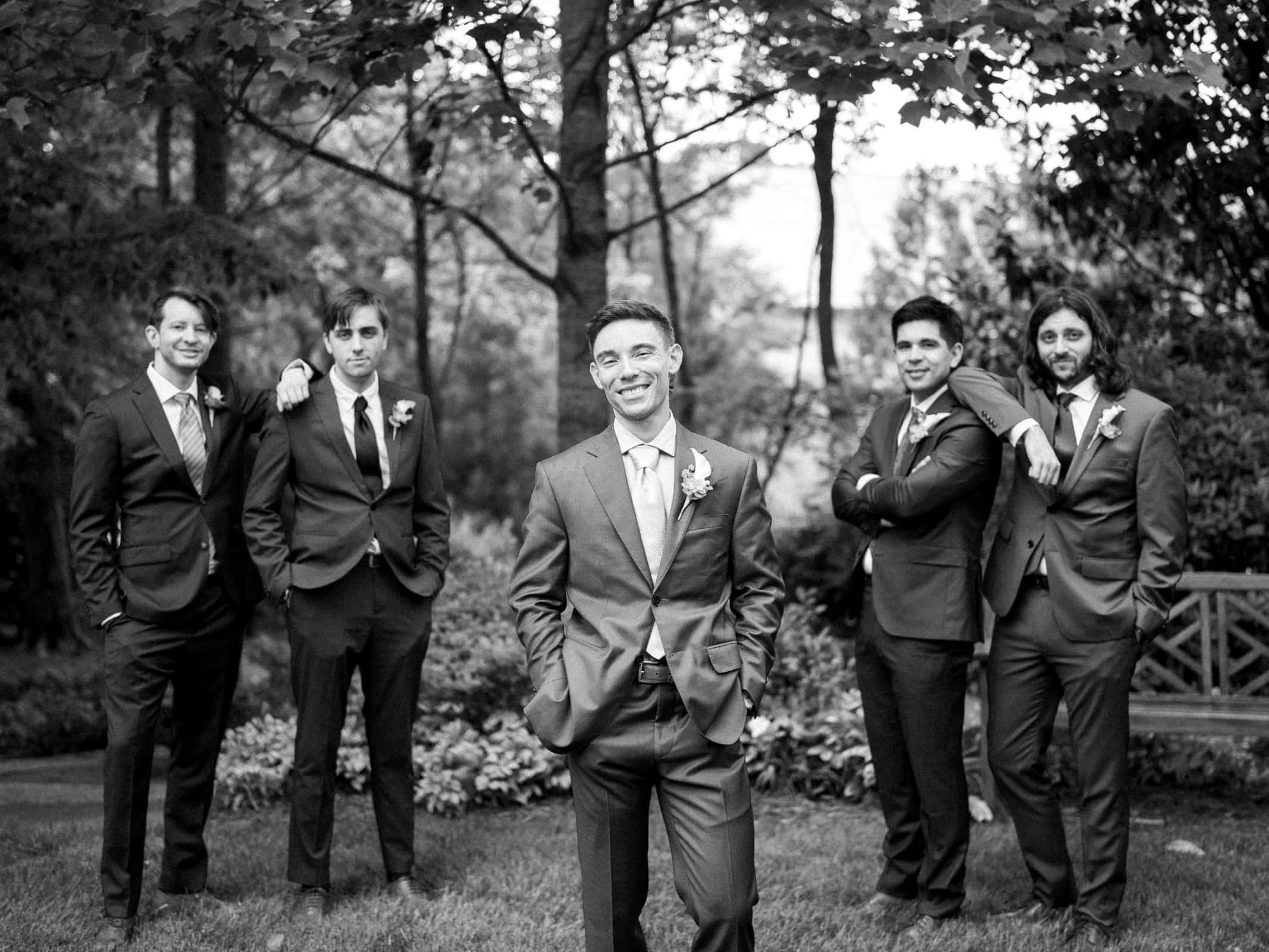 duke university wedding photography - sarah & case A black and white photo captures five men in suits standing casually on a lawn, reminiscent of classic Duke University charm. The central figure stands with hands in pockets, exuding elegance, while the others gather around. Trees and a bench add to the timeless wedding photography style.