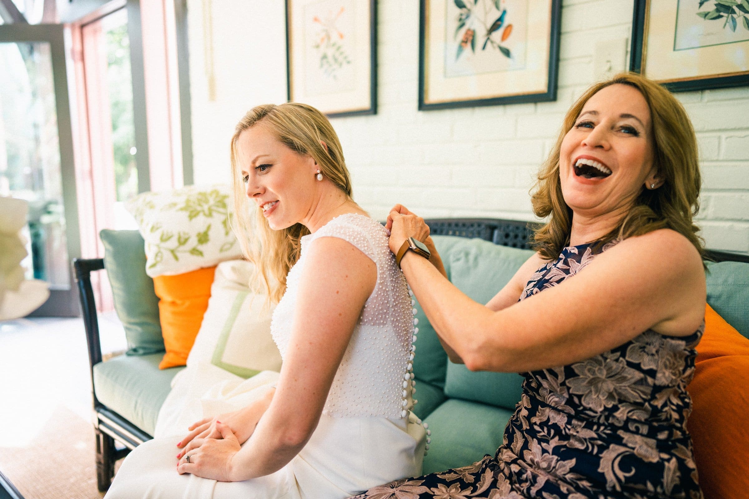 duke university wedding photography - sarah & case A woman in a white dress smiles as another woman, perhaps from Duke University, fastens the back of her gown. They are seated on a couch with colorful cushions, surrounded by framed artwork. Bright sunlight filters through a nearby window, capturing a perfect moment akin to wedding photography.