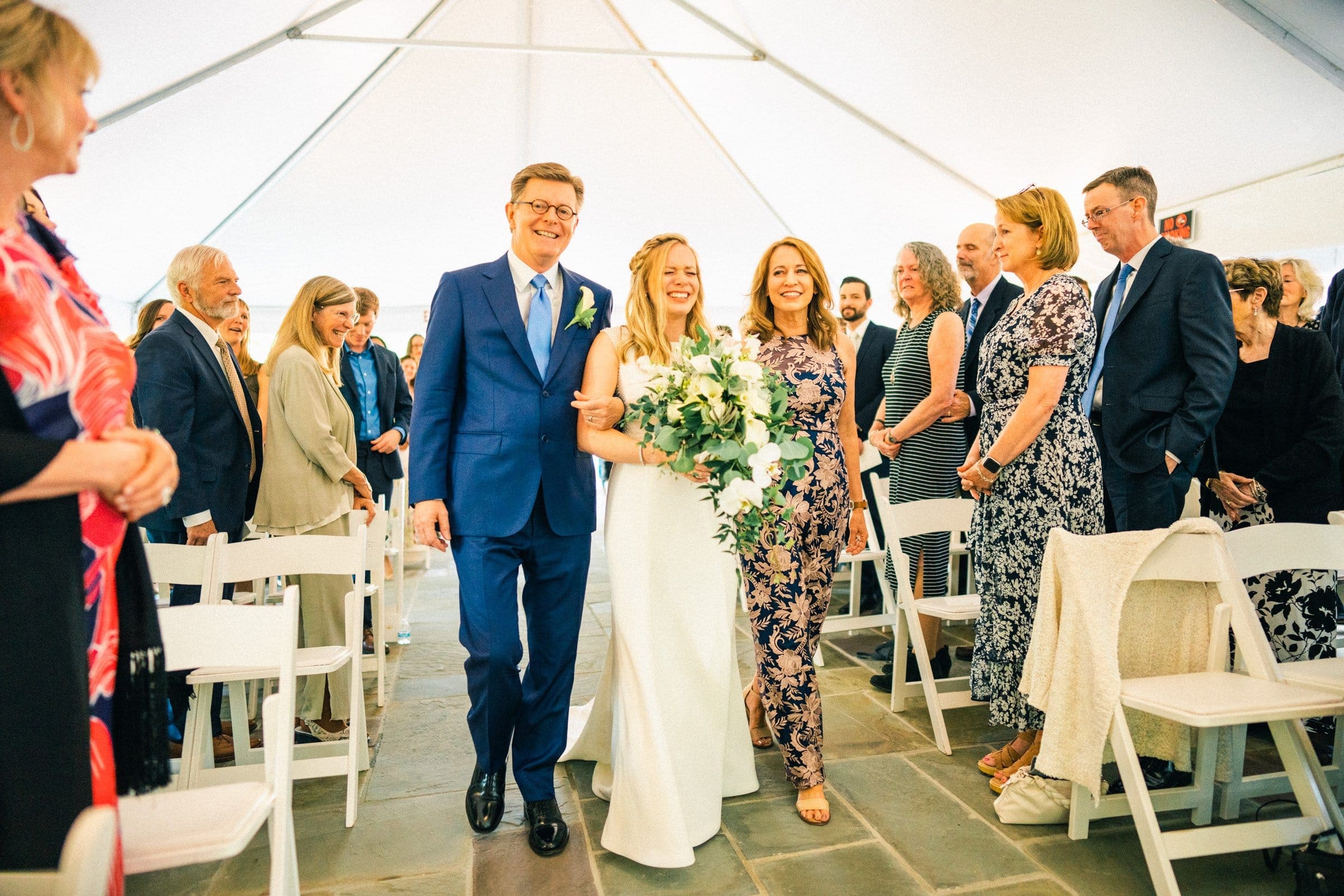 duke university wedding photography - sarah & case A bride walks down the aisle, smiling, holding a bouquet of white flowers. Accompanied by two people, she steps gracefully as guests in formal attire look on. Captured in stunning detail by Duke University wedding photography, the scene unfolds gracefully under a pristine white tent.