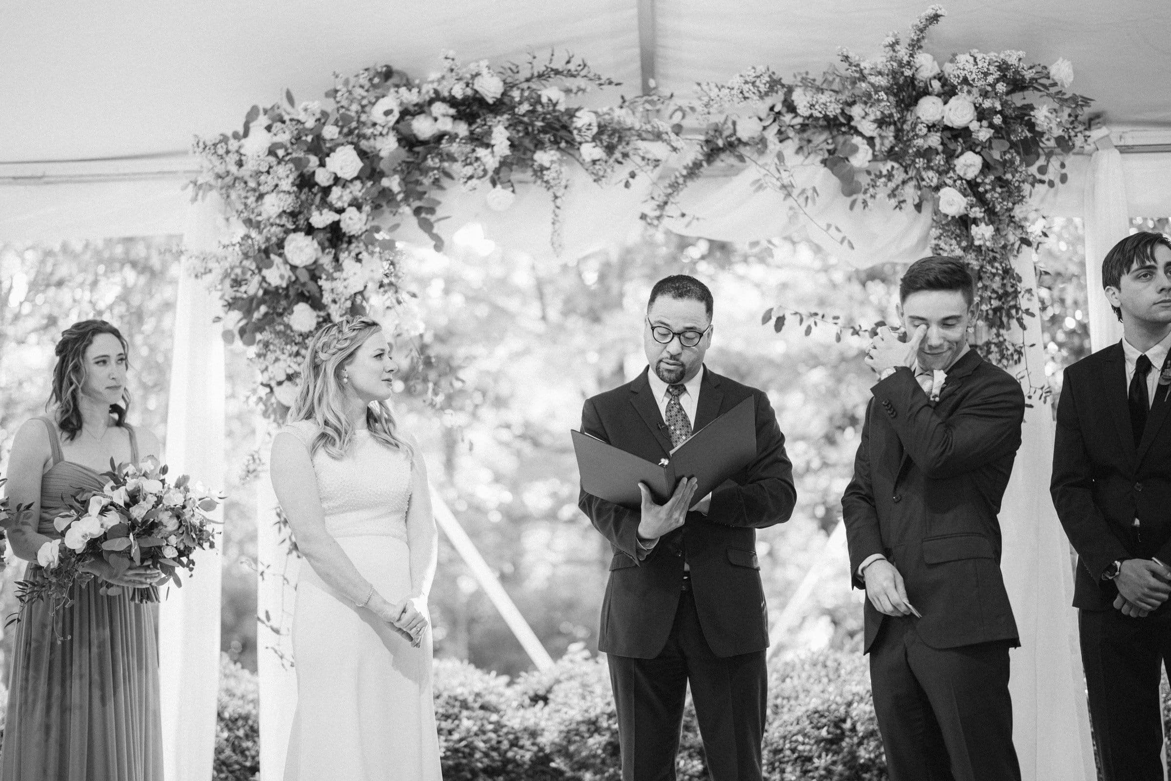 duke university wedding photography - sarah & case A black-and-white photo captures a wedding ceremony at Duke University under a floral arch. The bride beams as the groom appears emotional. An officiant reads from a book, while two attendants stand on either side, framing this touching moment in wedding photography.