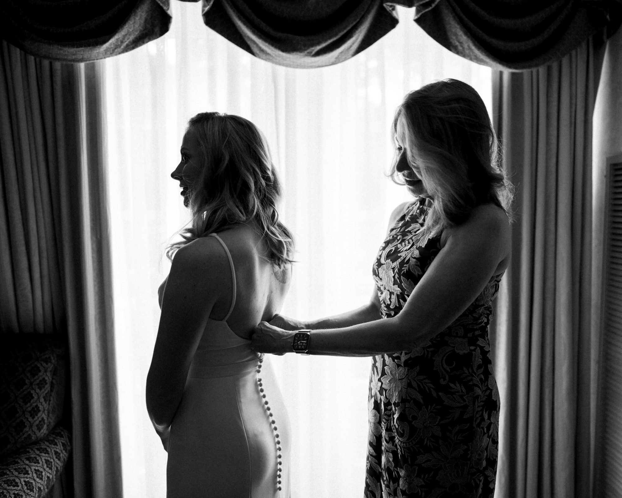 duke university wedding photography - sarah & case In a serene black-and-white scene reminiscent of Duke University wedding photography, a woman assists another with her dress in front of a bright window. Both have long hair and wear elegant attire, capturing an intimate moment filled with grace.
