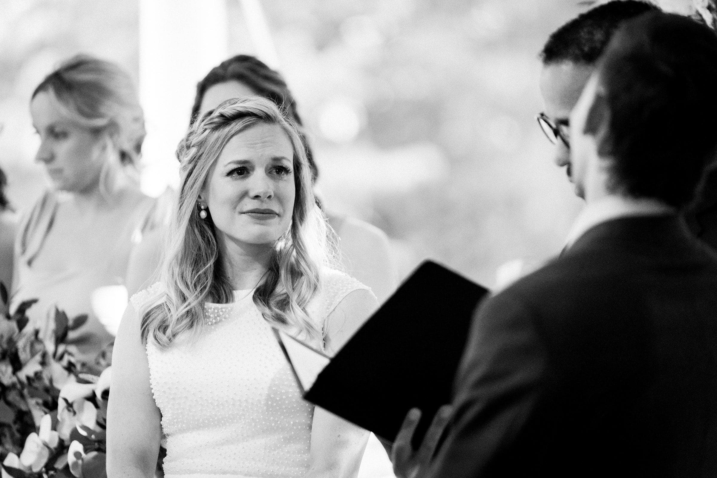 duke university wedding photography - sarah & case A black and white photo captures the Duke University wedding ceremony. A woman in a white dress stands, looking emotional, with bridesmaids behind her. A man in a suit, holding an open book, faces her as he speaks. The softly blurred background adds an intimate touch.