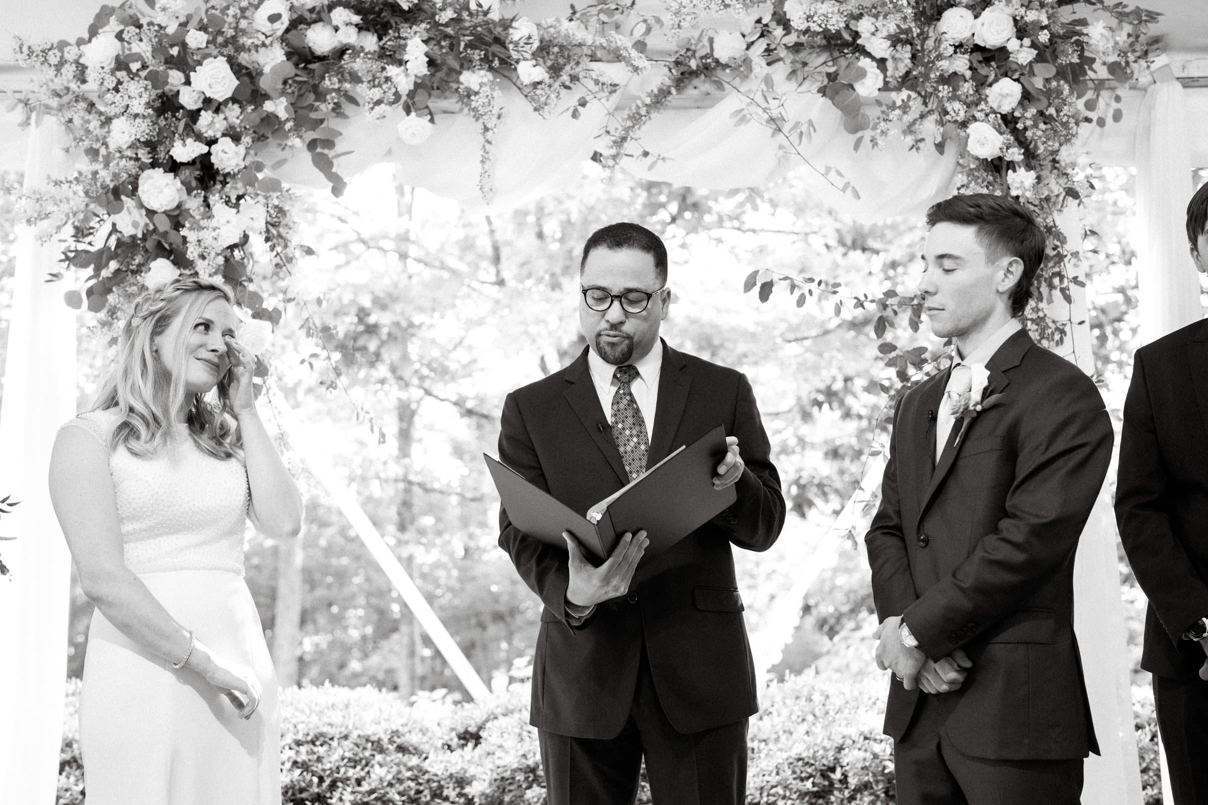 duke university wedding photography - sarah & case A black and white photo captures a Duke University wedding ceremony. A woman in a white dress wipes a tear beside a man in a suit, as the officiant reads from a book. Floral decorations are visible above them, creating an elegant backdrop for this poignant moment.