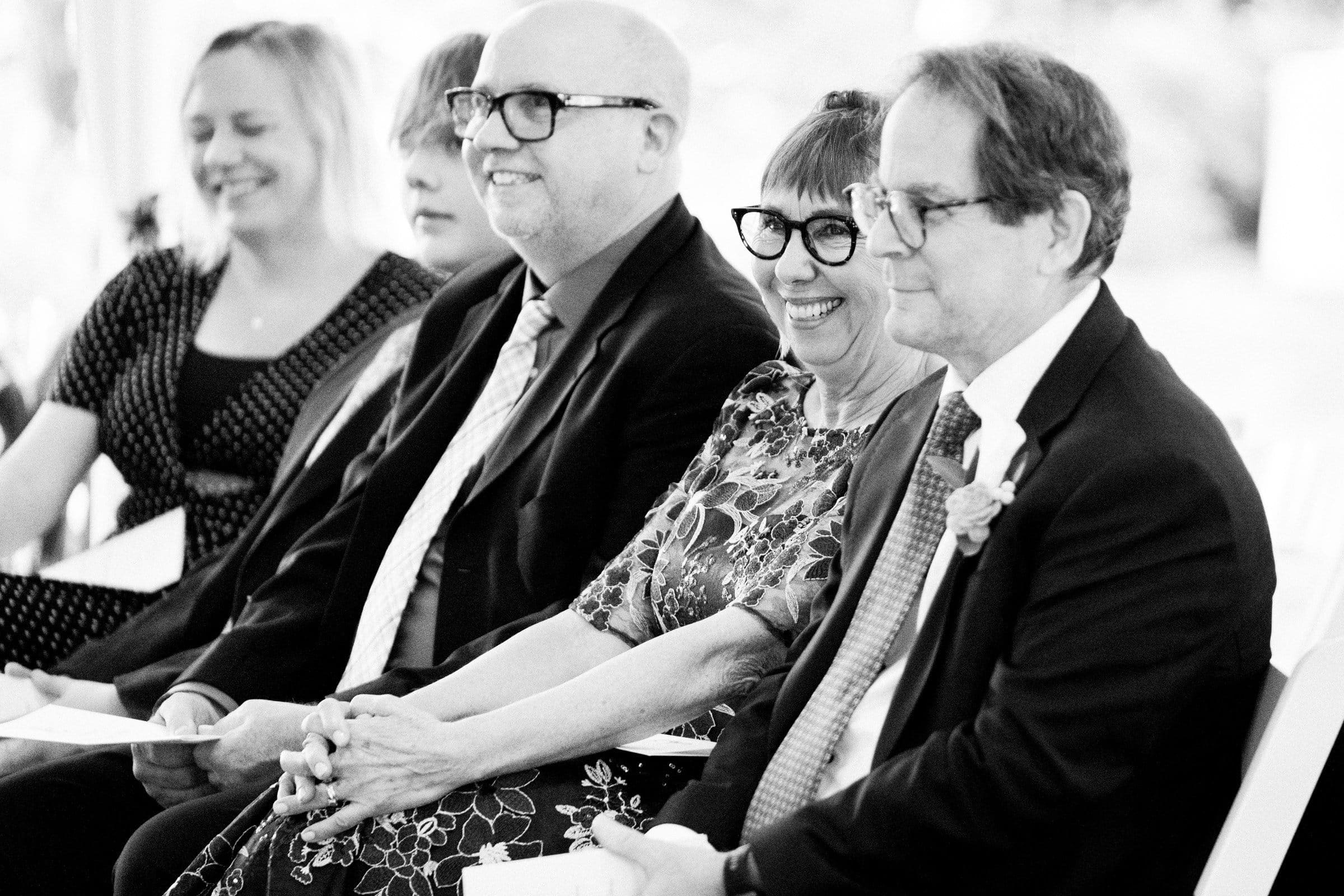 duke university wedding photography - sarah & case A black and white photo captures a group of people sitting in a row at what seems to be a formal event, perhaps related to Duke University. They are smiling and engaged, with the men in suits and the woman wearing glasses and a floral dress.