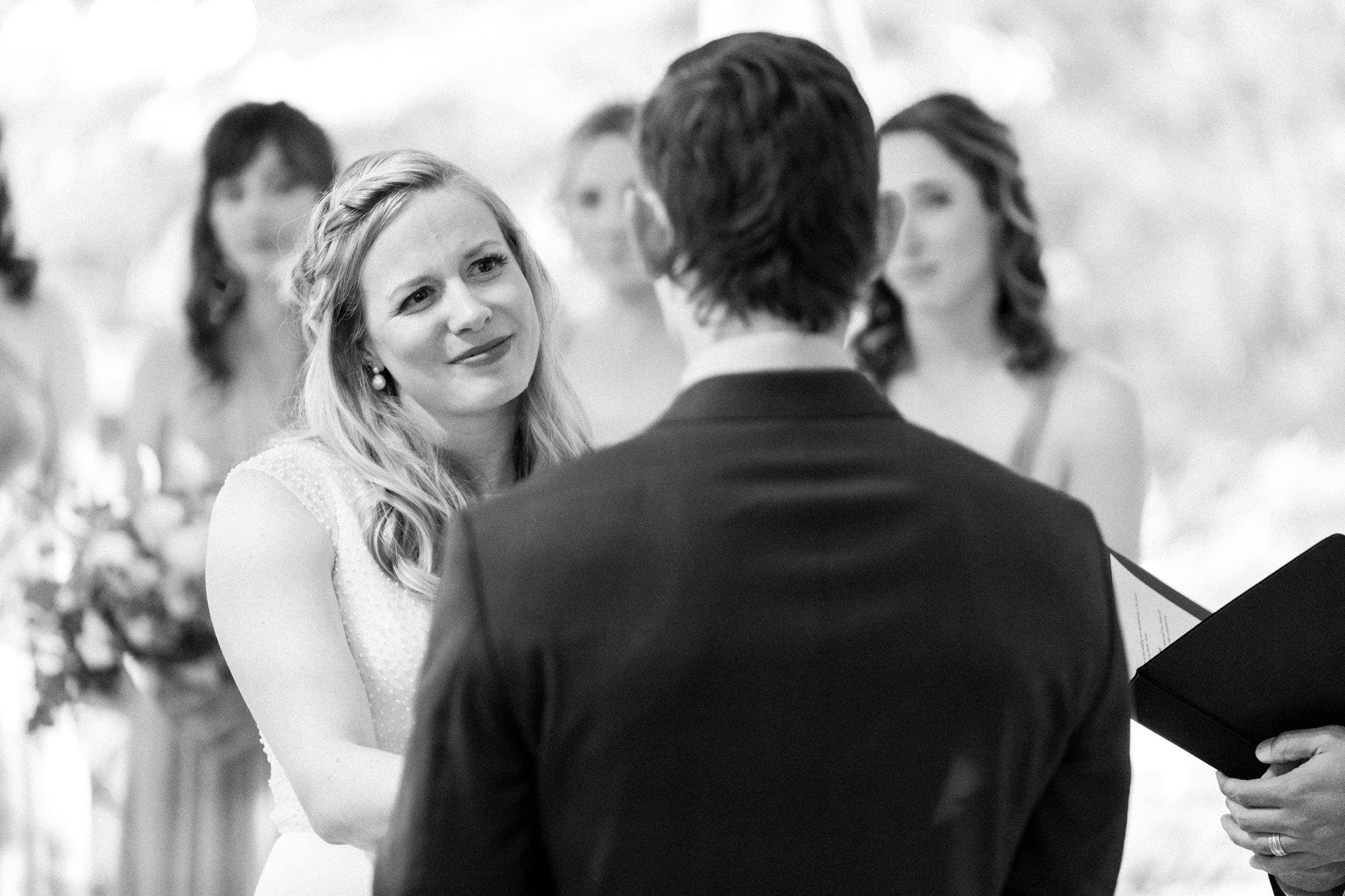 duke university wedding photography - sarah & case A black and white photo of a Duke University wedding ceremony captures a bride with braided hair looking lovingly at the groom, who has his back to the camera. Bridesmaids and an officiant holding documents are elegantly positioned in the background, enhancing this timeless wedding photography moment.