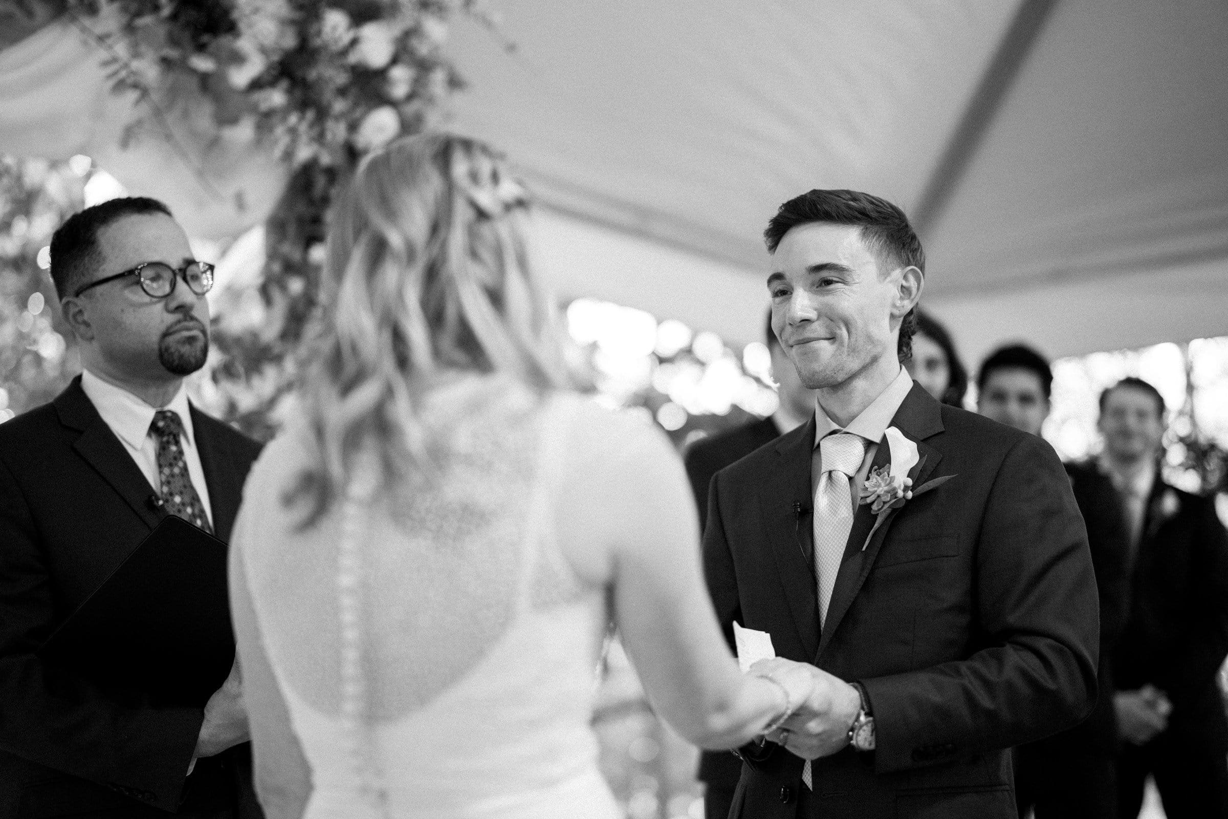 duke university wedding photography - sarah & case A black and white photo captures a Duke University wedding ceremony. The groom in a suit smiles as he holds the hands of the bride, whose back is to the camera. A man in glasses stands in the background, likely officiating, under a tent adorned with floral decorations.