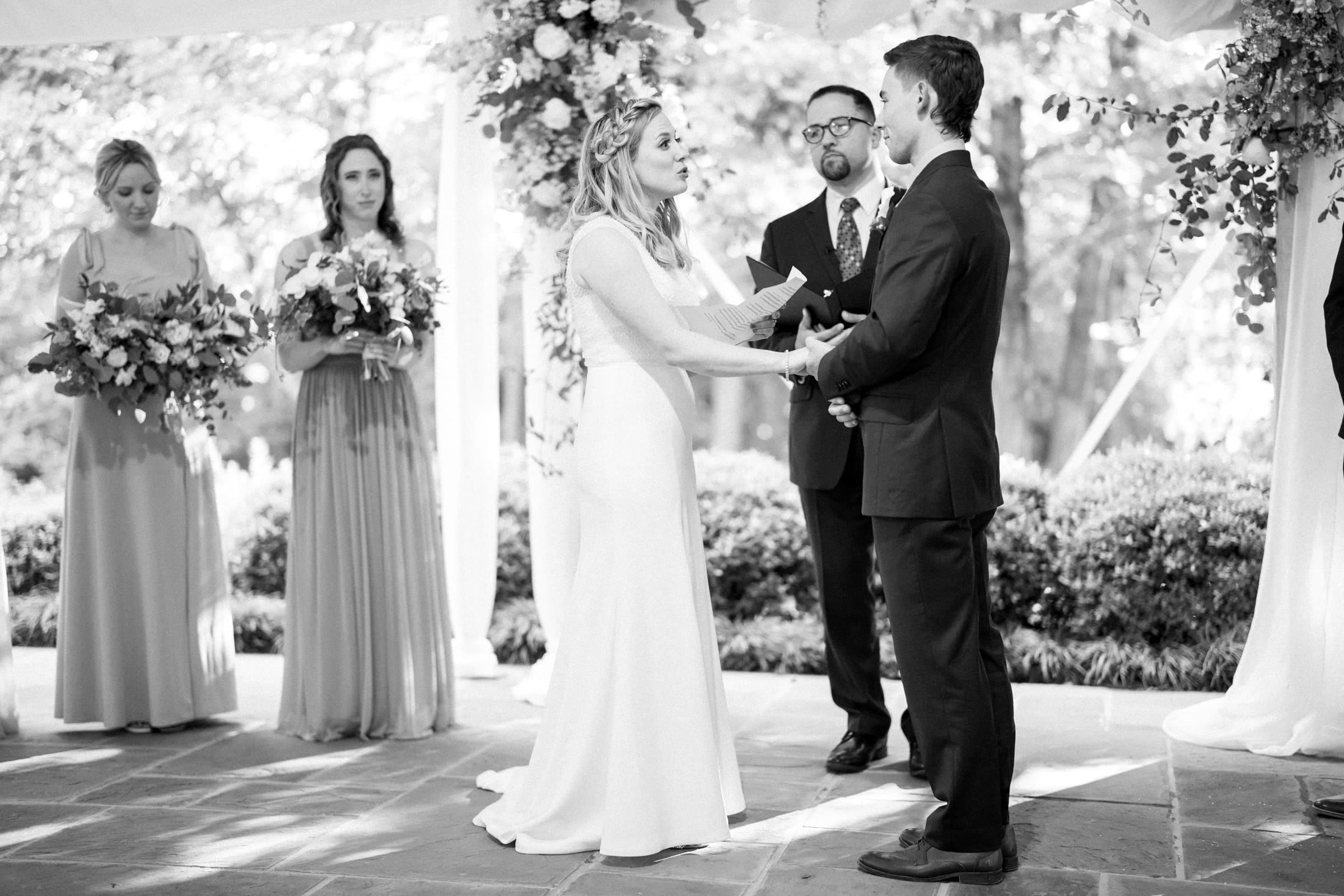 duke university wedding photography - sarah & case A bride and groom stand facing each other, holding hands under a beautifully decorated arch during a wedding ceremony. Bridesmaids with vibrant bouquets grace the left side as the officiant presides. This enchanting moment is captured amidst lush greenery by Duke University wedding photography.