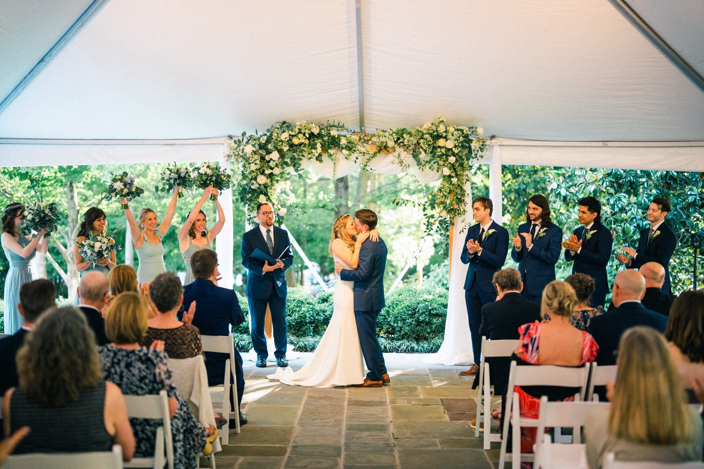 duke university wedding photography - sarah & case A bride and groom share a kiss under a floral arch at an outdoor wedding ceremony, beautifully captured by Duke University wedding photography. They are surrounded by the bridal party—bridesmaids in green dresses and groomsmen in dark suits—while guests look on, enchanted by the moment.