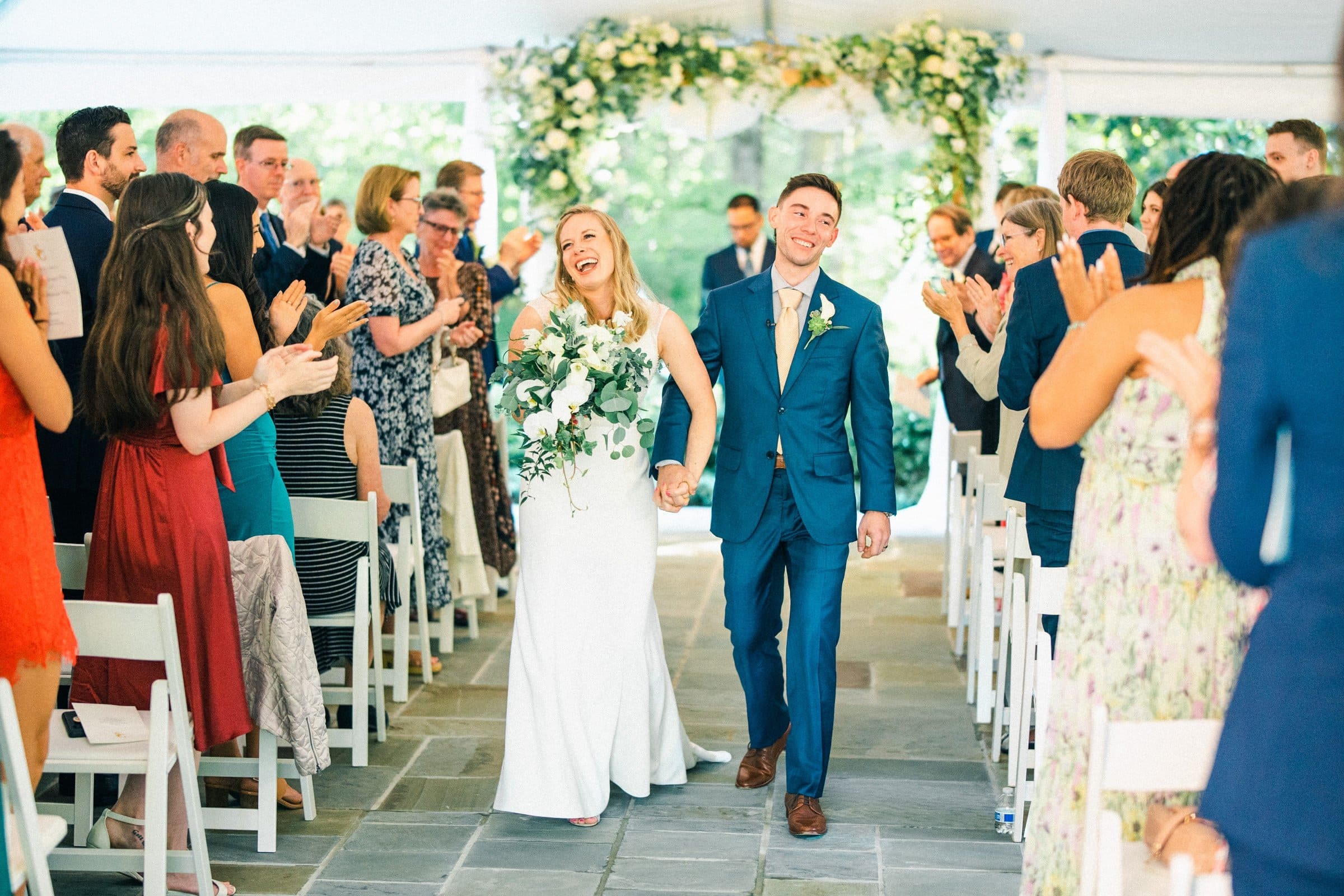 duke university wedding photography - sarah & case A bride and groom walk down the aisle at Duke University, smiling broadly amid clapping guests. The bride holds a bouquet and wears a white dress, while the groom is in a blue suit. The venue's elegant flowers add to the charming wedding photography backdrop.