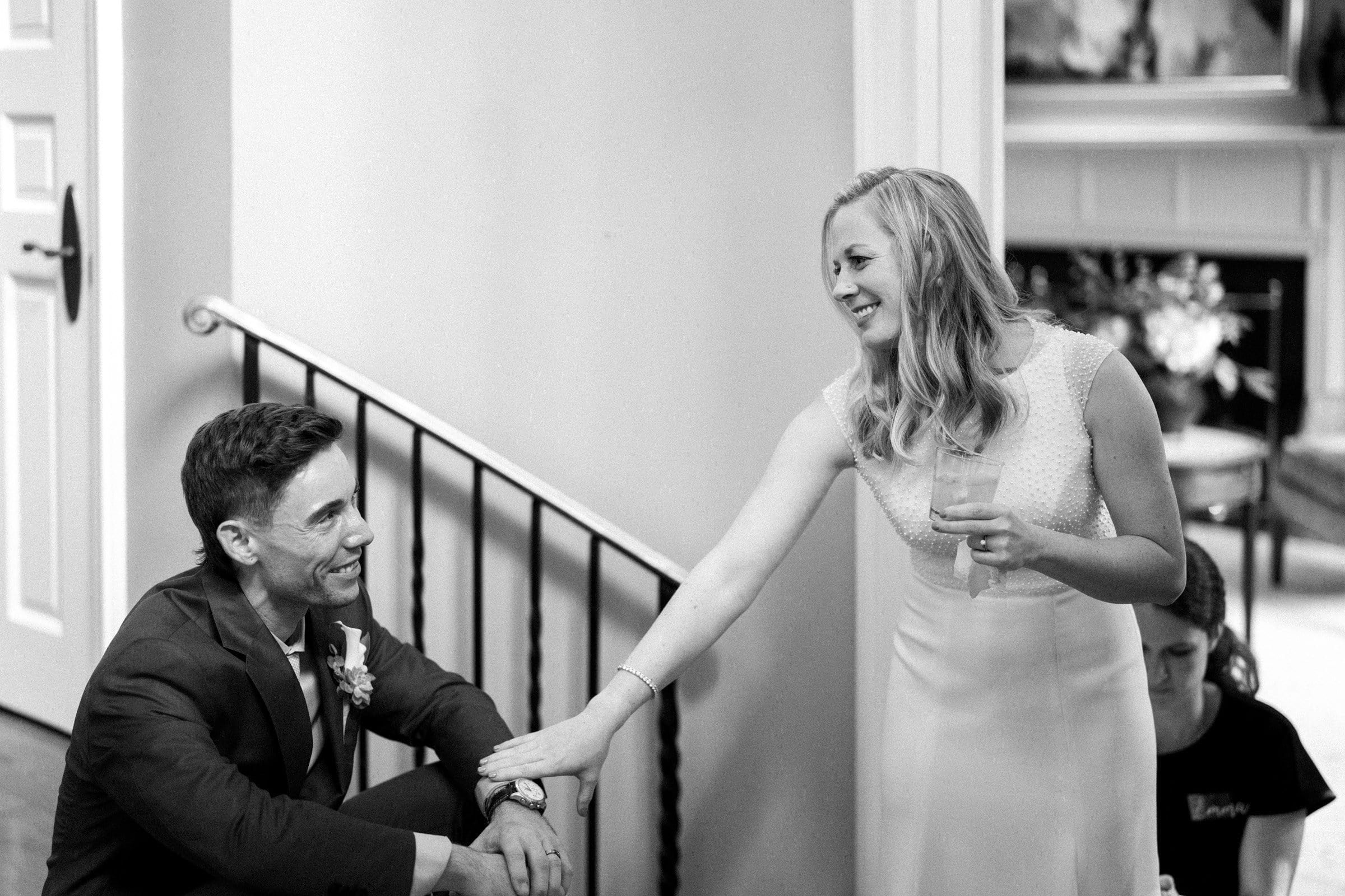 duke university wedding photography - sarah & case A black and white photo captures a smiling couple indoors, epitomizing classic Duke University wedding photography. The woman, in a light-colored dress, holds a drink and gently touches the man’s hand. Seated on a staircase, he wears a suit with a boutonniere, looking up at her adoringly.