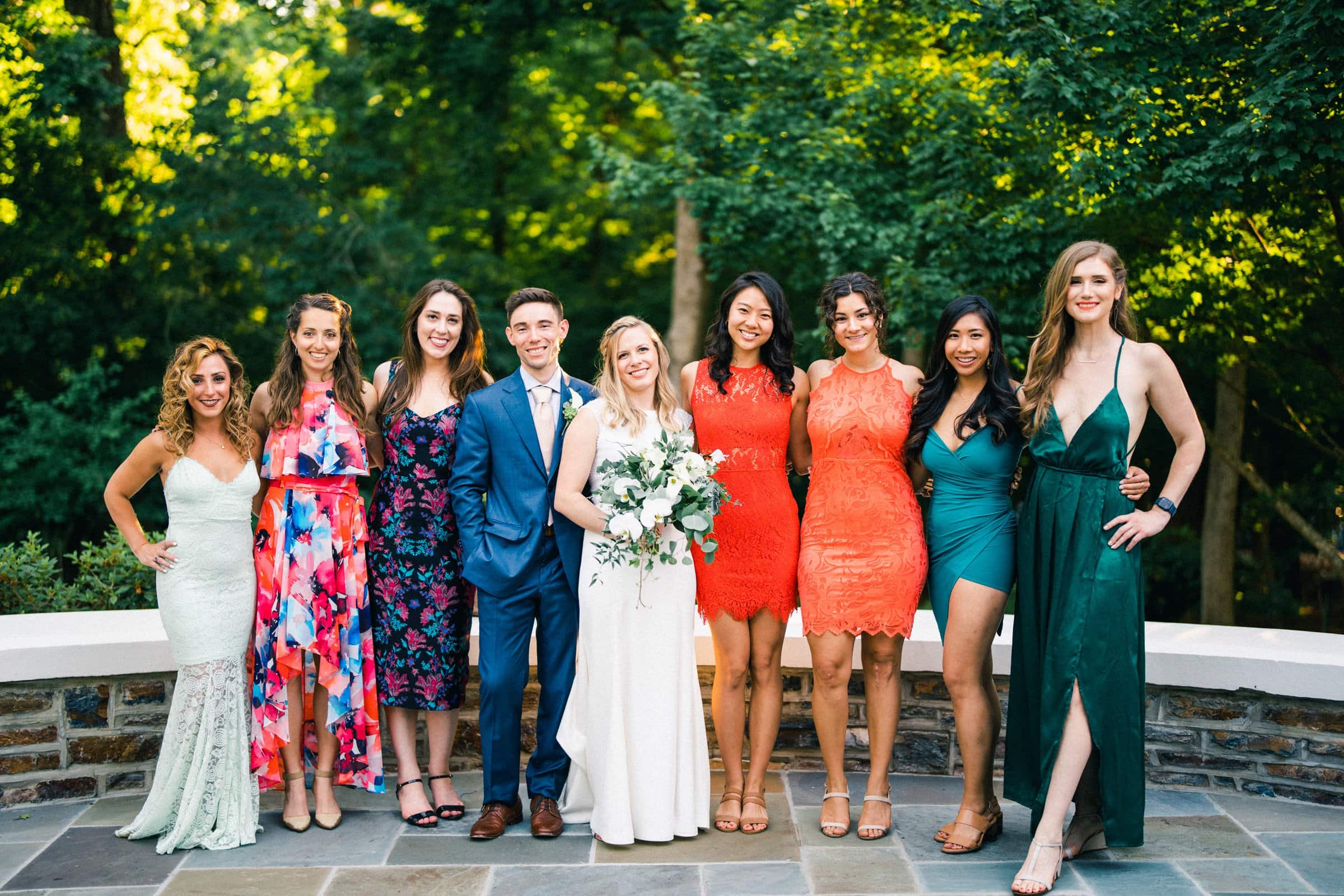 duke university wedding photography - sarah & case A group of people dressed in formal attire pose together outdoors in a classic display of Duke University wedding photography. The bride stands in the center holding a bouquet, surrounded by bridesmaids and groomsmen in colorful dresses and suits, with lush greenery providing the perfect backdrop.