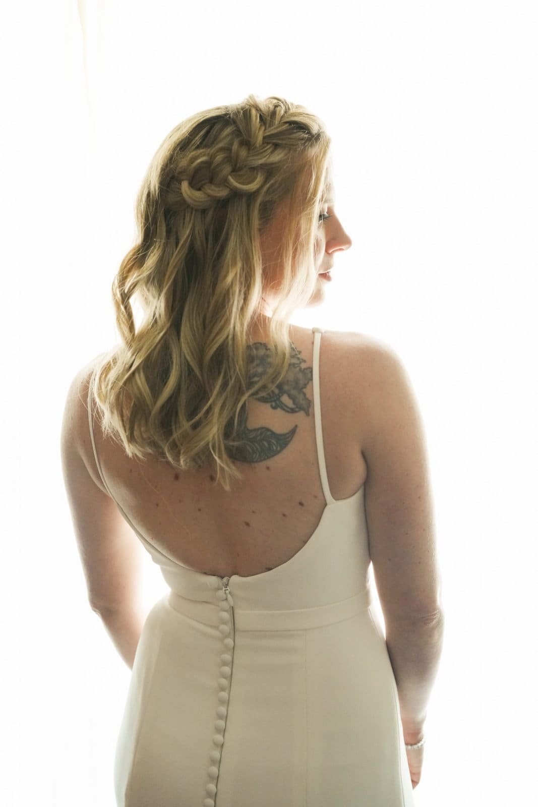 duke university wedding photography - sarah & case A bride in a white dress stands with her back to the camera, showing off a braided hairstyle and a tattoo on her upper back. Captured with Duke University wedding photography expertise, the bright background highlights her hair and dress details beautifully.