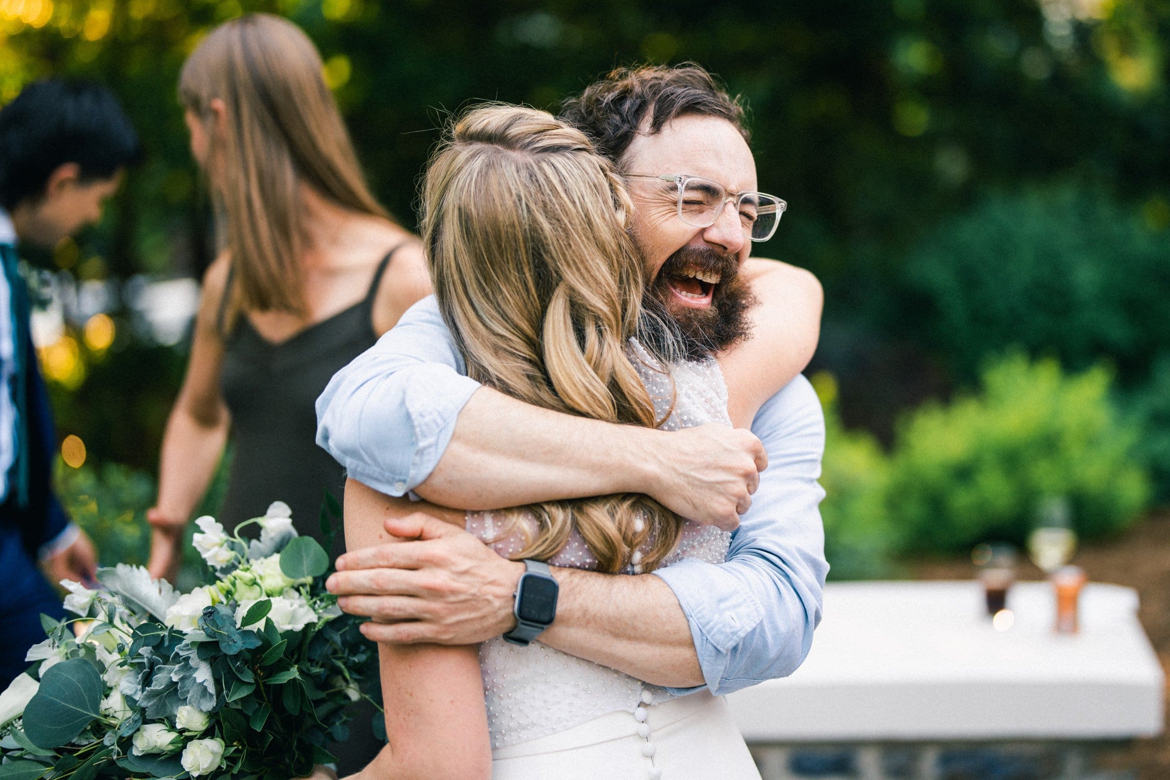 duke university wedding photography - sarah & case A joyful bearded man in glasses and a light blue shirt hugs a woman with long blonde hair in a white dress, her bouquet of green and white flowers complementing the scene. It's as if Duke University wedding photography captured this moment amid blurred greenery and smiling faces in the background.