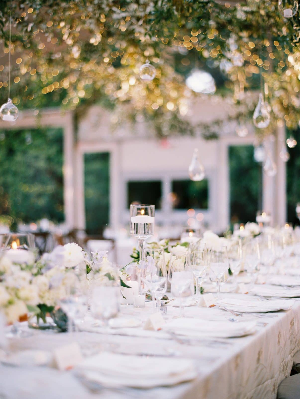 duke university wedding photography - sarah & case A long, elegantly set dining table adorned with white flowers and candles exudes romance under soft fairy lights. This enchanting outdoor garden setting is beautifully captured by Duke University wedding photography, with glasses and tableware neatly arranged for the perfect celebration.