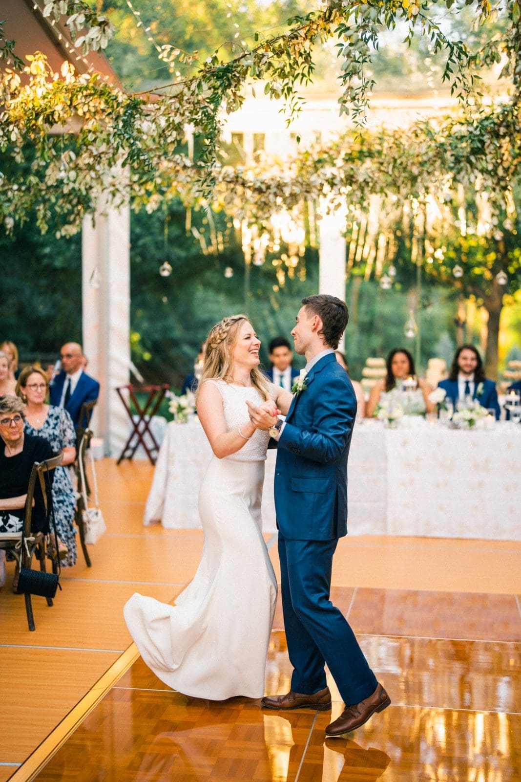 duke university wedding photography - sarah & case A bride in a white dress and a groom in a blue suit dance on a wooden floor under a canopy adorned with lights and greenery, capturing the essence of Duke University wedding photography. Guests seated at tables in the background watch and smile.