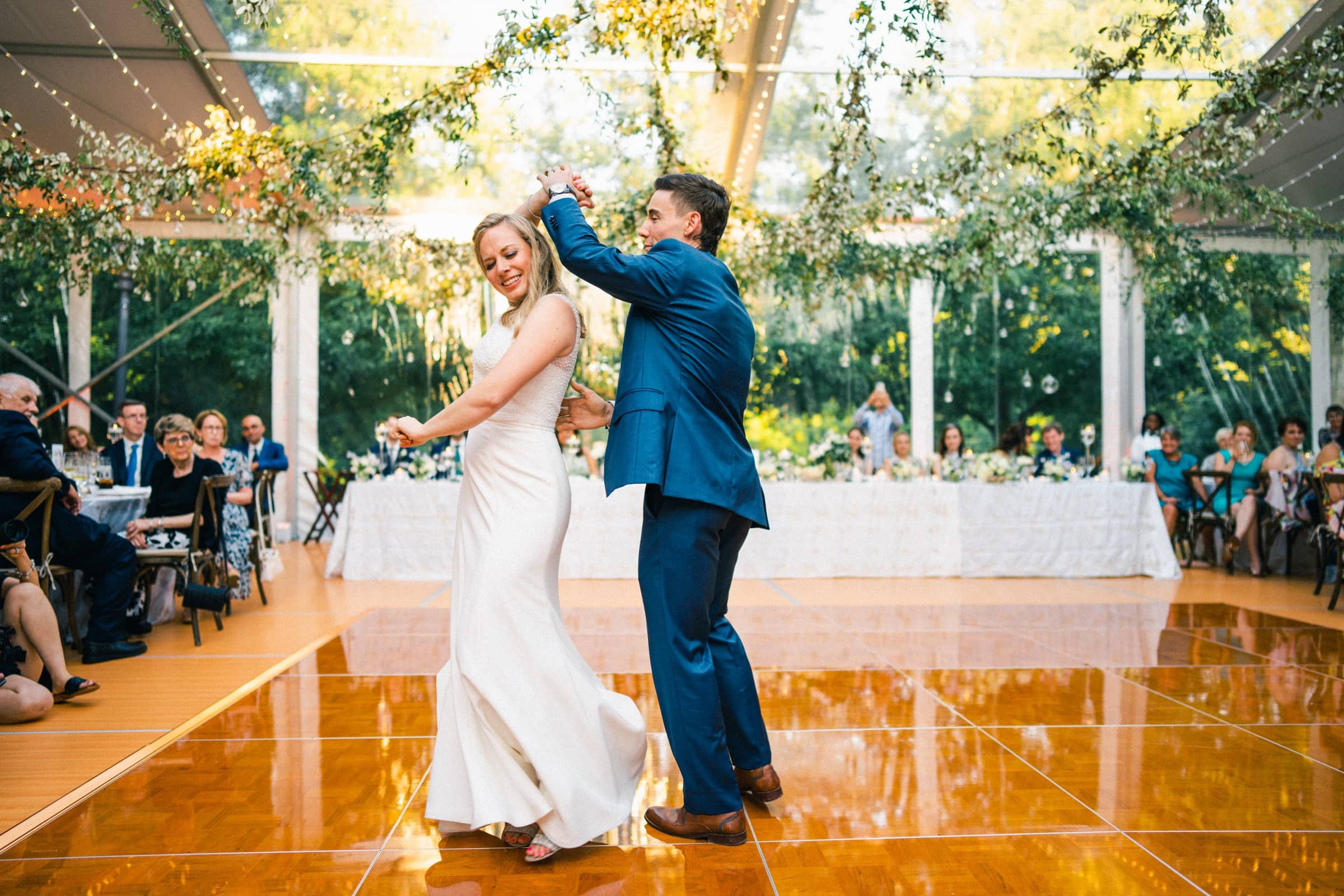 duke university wedding photography - sarah & case A bride and groom dance joyfully on a polished wooden floor under a tent adorned with greenery and lights. Captured by Duke University wedding photography, guests are seated at tables, watching the couple with smiles. Sunlight streams through the transparent roof, creating a warm ambiance.