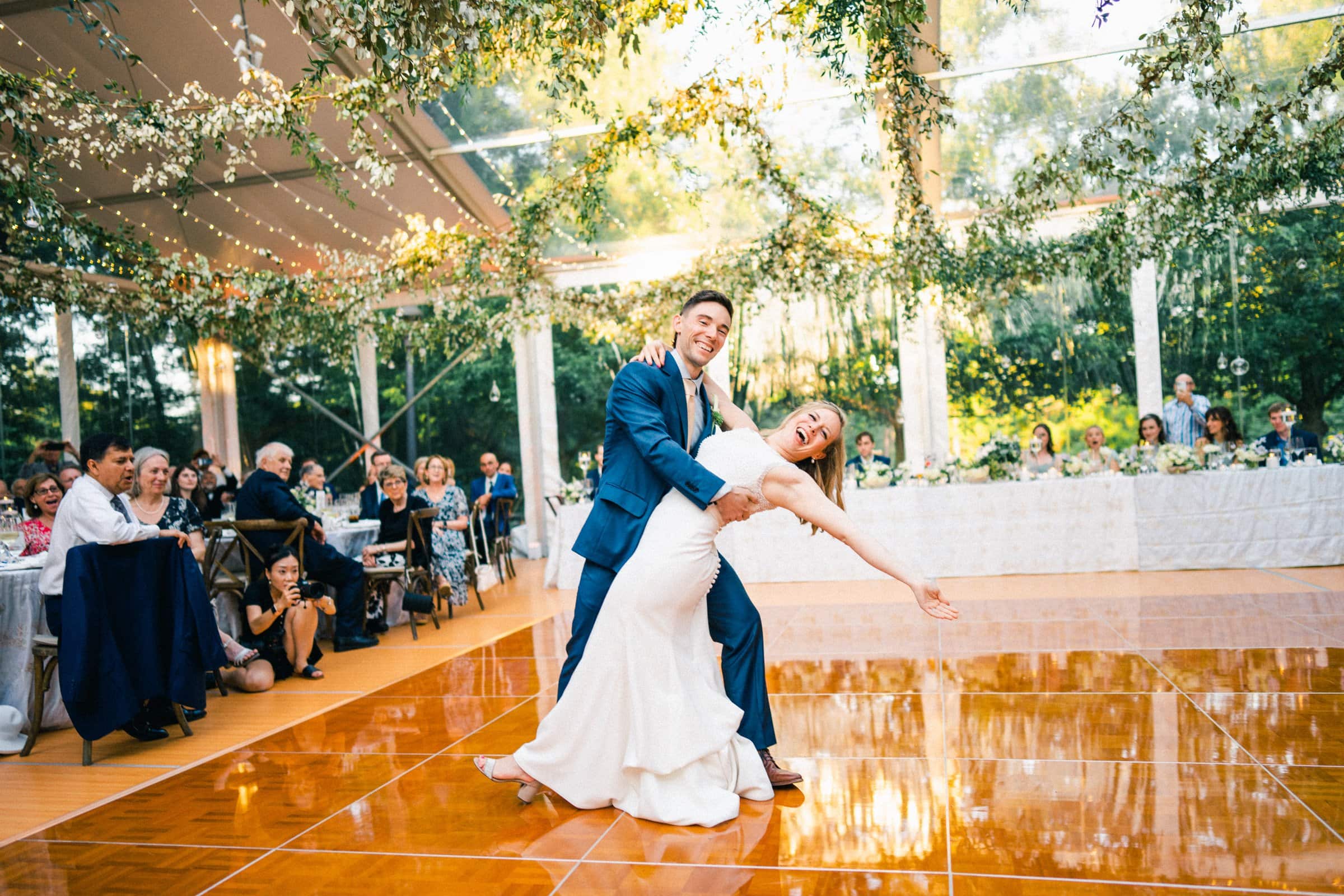 duke university wedding photography - sarah & case A couple in formal attire shares a joyful dance on a wooden floor under a tent adorned with greenery and fairy lights, captured beautifully by Duke University wedding photography. Guests seated at tables in the background watch and smile, enhancing the festive and romantic atmosphere.