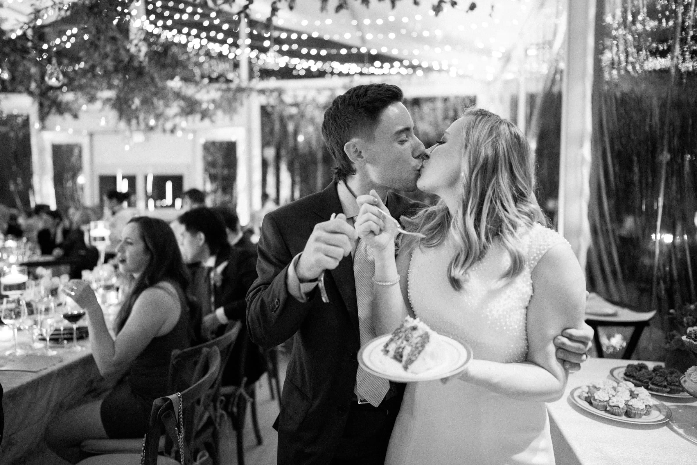 duke university wedding photography - sarah & case At a beautifully adorned wedding reception, a couple shares a tender kiss while holding a plate with a piece of cake. The room glows with string lights, and guests relish the celebration in the background, captured perfectly by Duke University wedding photography.