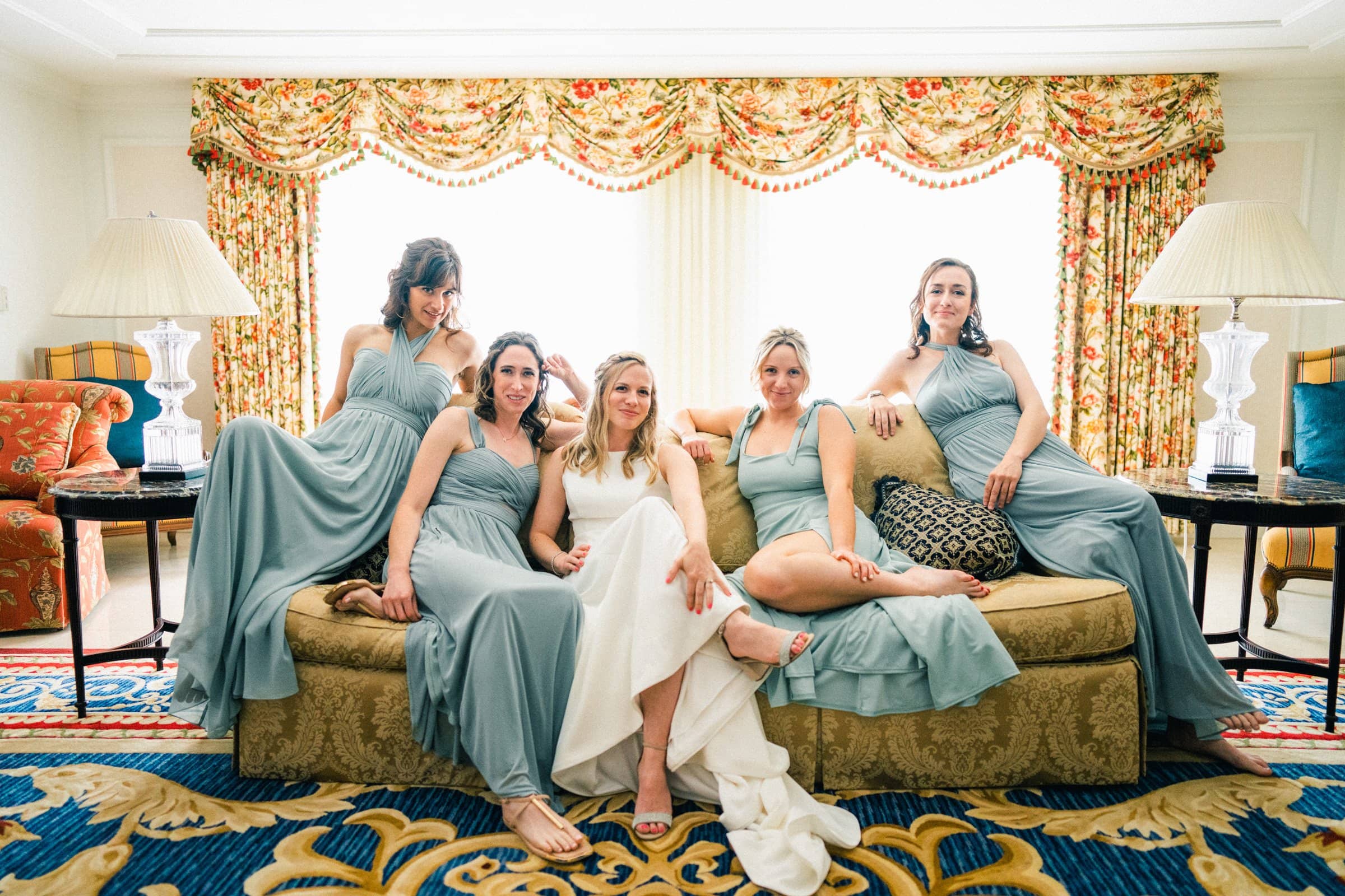 duke university wedding photography - sarah & case Five women in light blue dresses sit and lean on a sofa in a well-decorated room with ornate curtains and lamps. They pose together comfortably, smiling and relaxed, capturing the joy of the moment akin to Duke University's famed wedding photography, in a brightly lit, elegant setting.