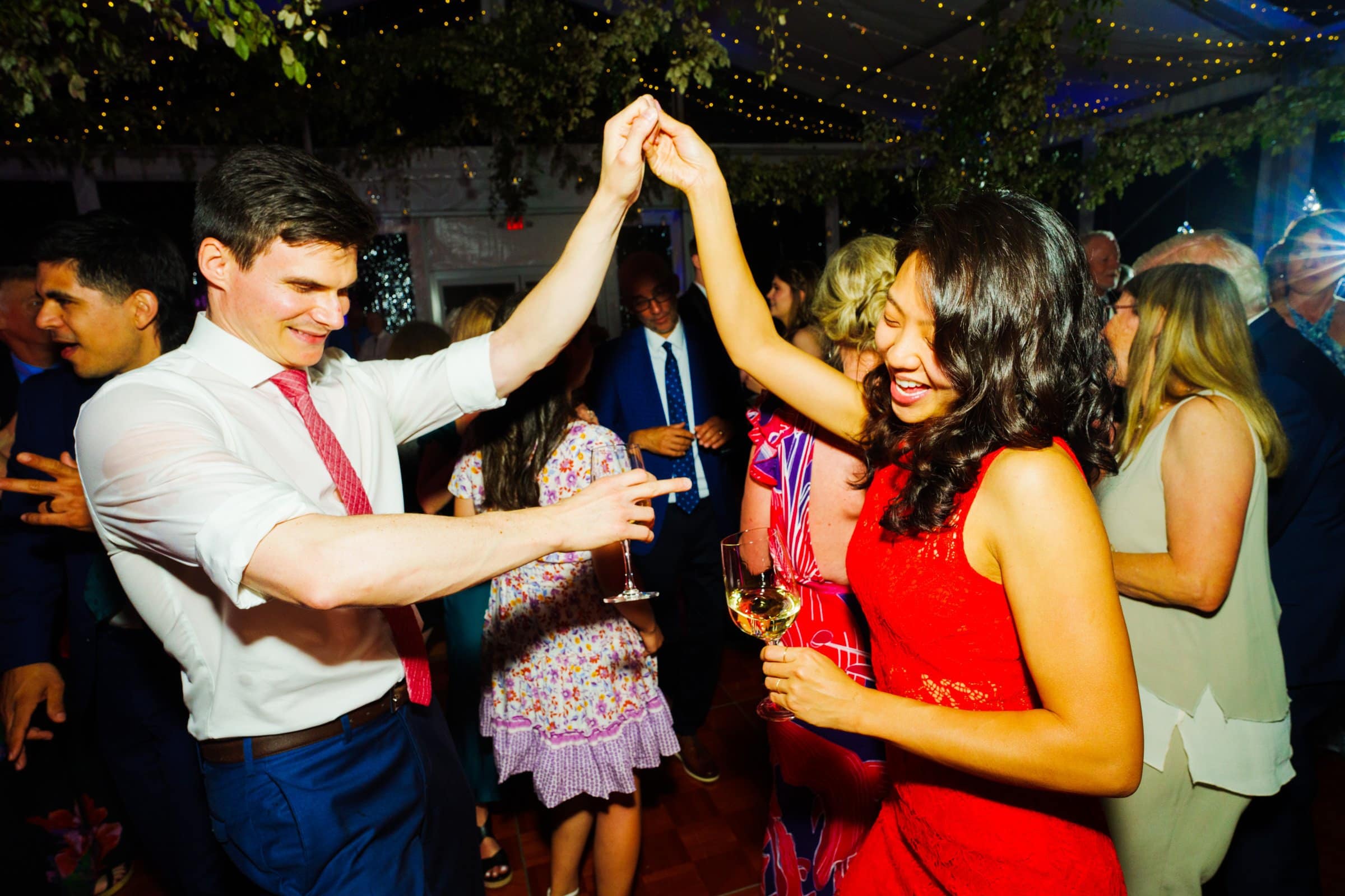duke university wedding photography - sarah & case A couple is dancing joyfully under twinkling lights at a lively Duke University event. The man, wearing a white shirt and red tie, and the woman, in a red dress, hold hands. Guests around them are mingling and enjoying the festive atmosphere, perfectly captured in stunning wedding photography.