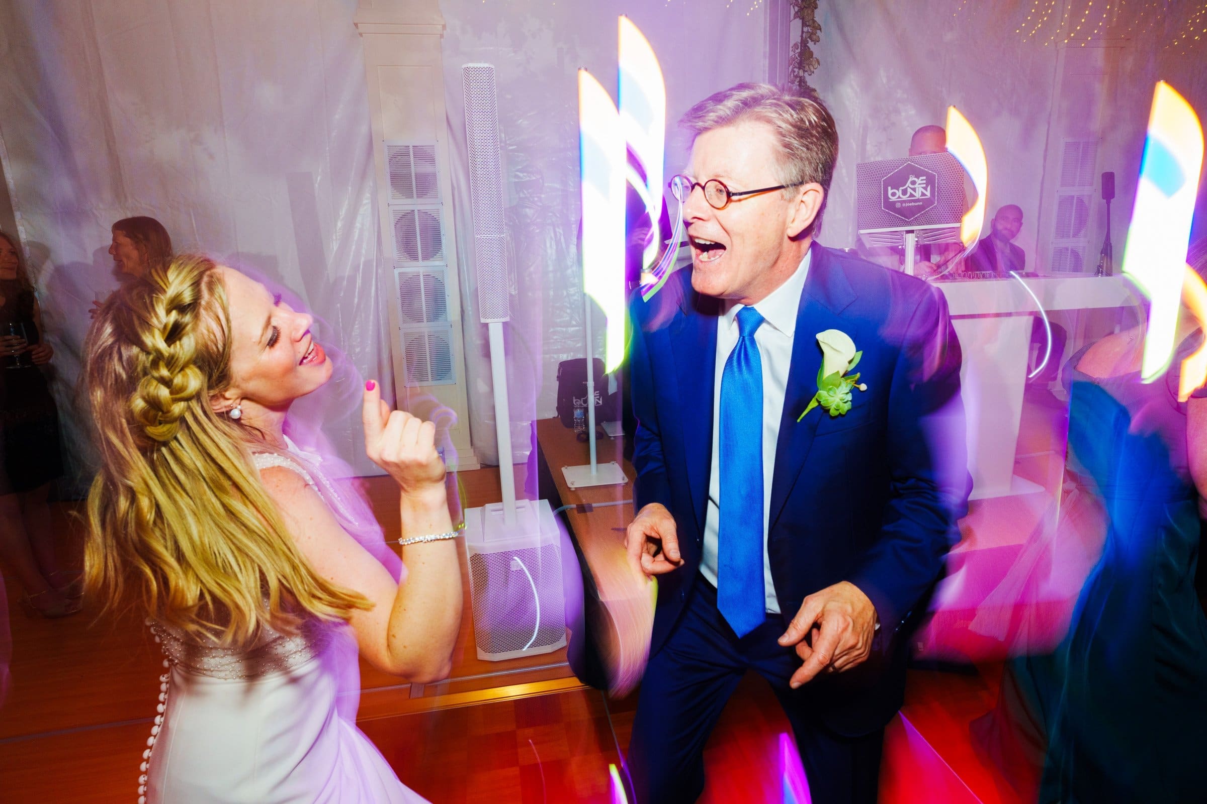 duke university wedding photography - sarah & case A woman in a white dress and a man in a blue suit dance energetically at an indoor event, captured in stunning Duke University wedding photography. Colorful light streaks and a DJ booth create an electrifying party atmosphere.