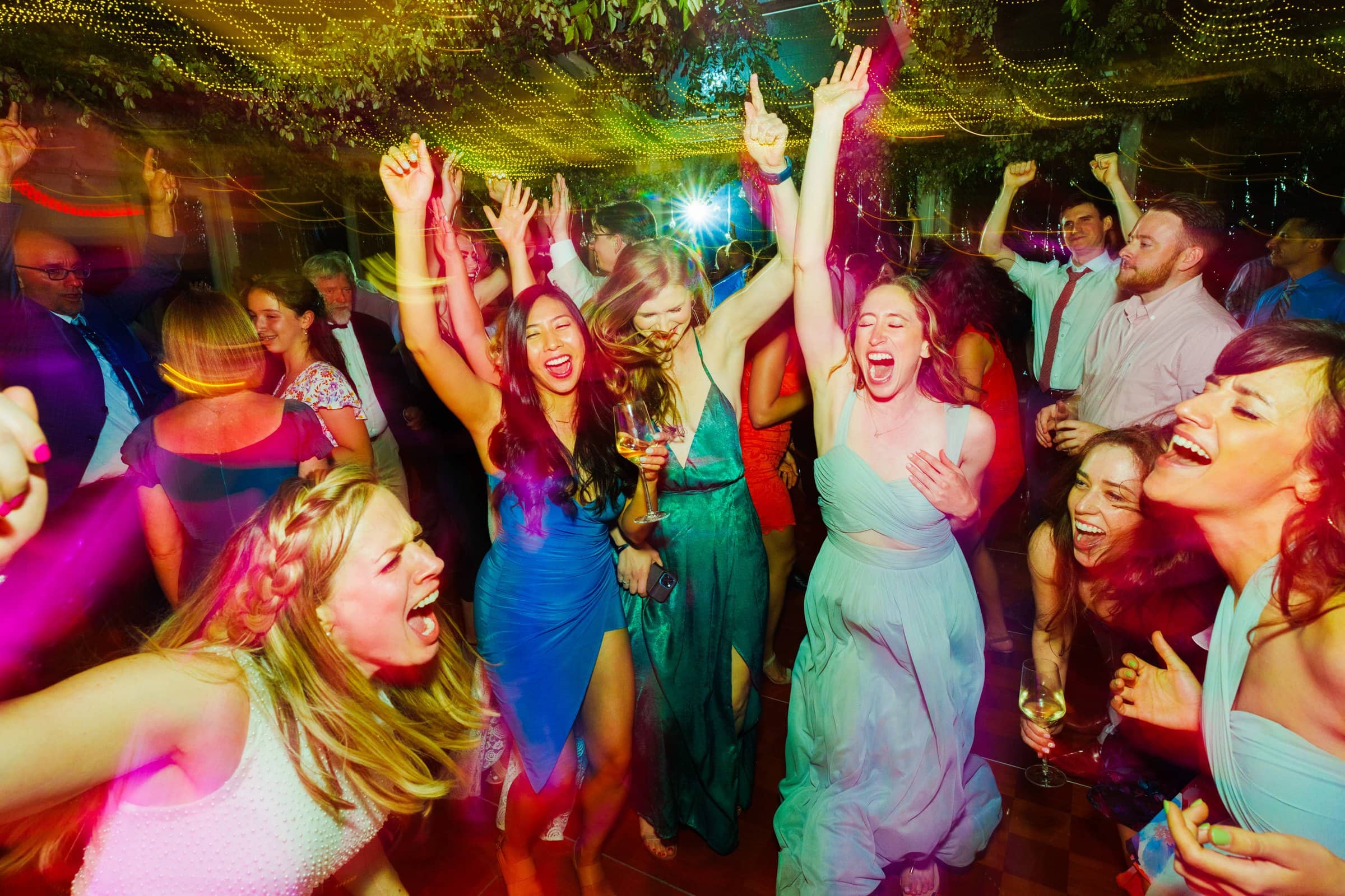 duke university wedding photography - sarah & case A lively group of people dancing with excitement at a Duke University party. The scene is vibrant, with colorful lights and partygoers in elegant attire celebrating, some with raised hands and glasses—capturing a moment worthy of wedding photography.