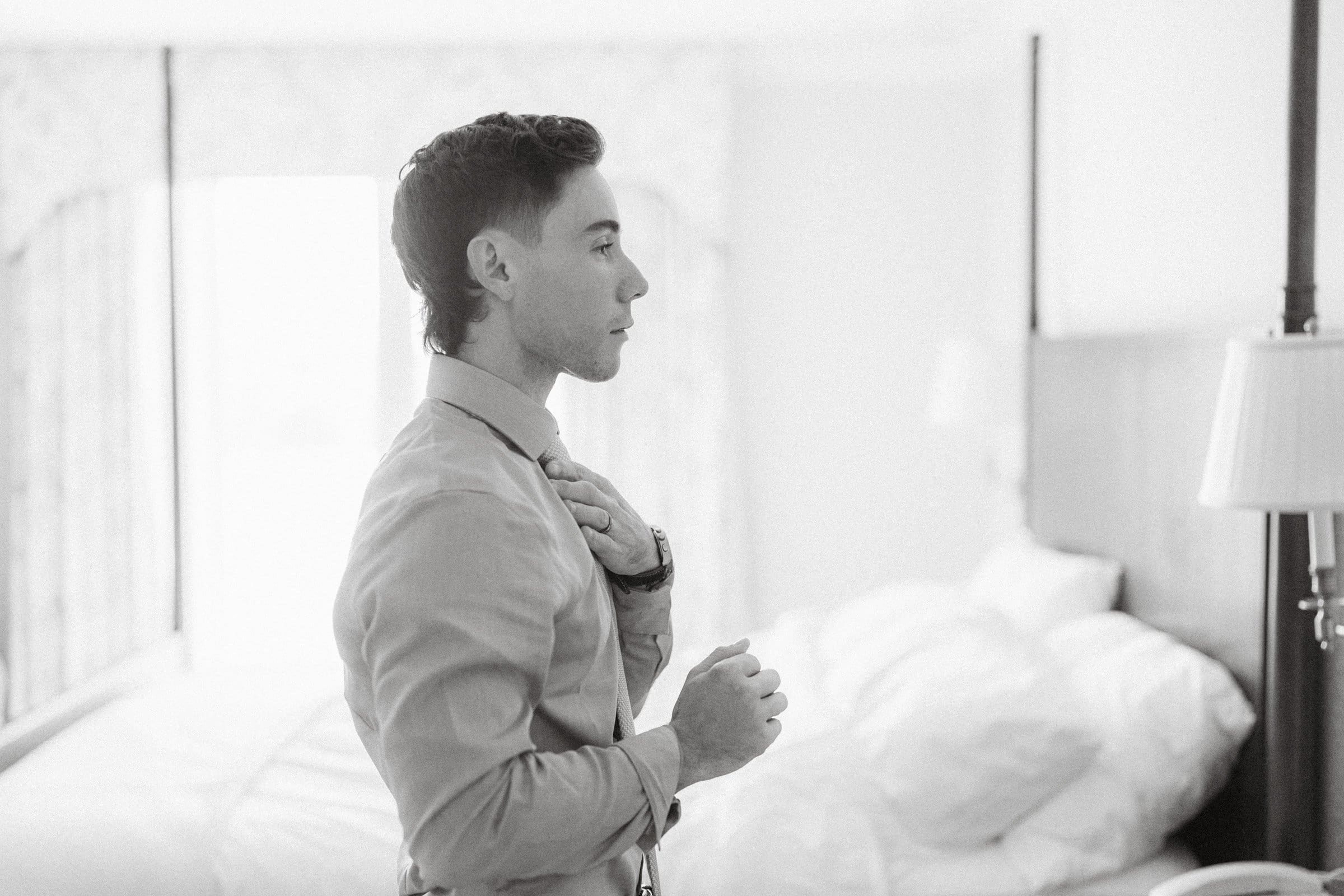 duke university wedding photography - sarah & case A black and white photo captures a man in a dress shirt adjusting his tie, reminiscent of Duke University alumni preparing for a celebration, standing in a bedroom with an unmade bed and a lamp gently illuminating the background.