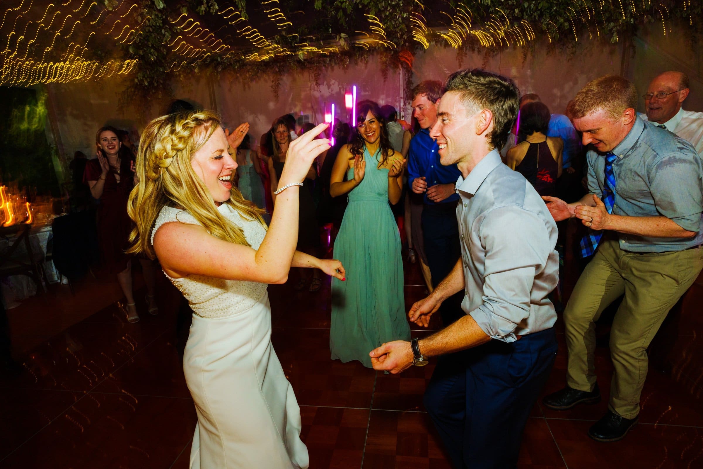 duke university wedding photography - sarah & case A joyful couple dances under string lights at a lively party, captured by a talented Duke University wedding photographer. The woman, in a white dress, holds a glowing pink stick, while the man wears a light shirt and dark pants. Guests in colorful attire cheer in the background.