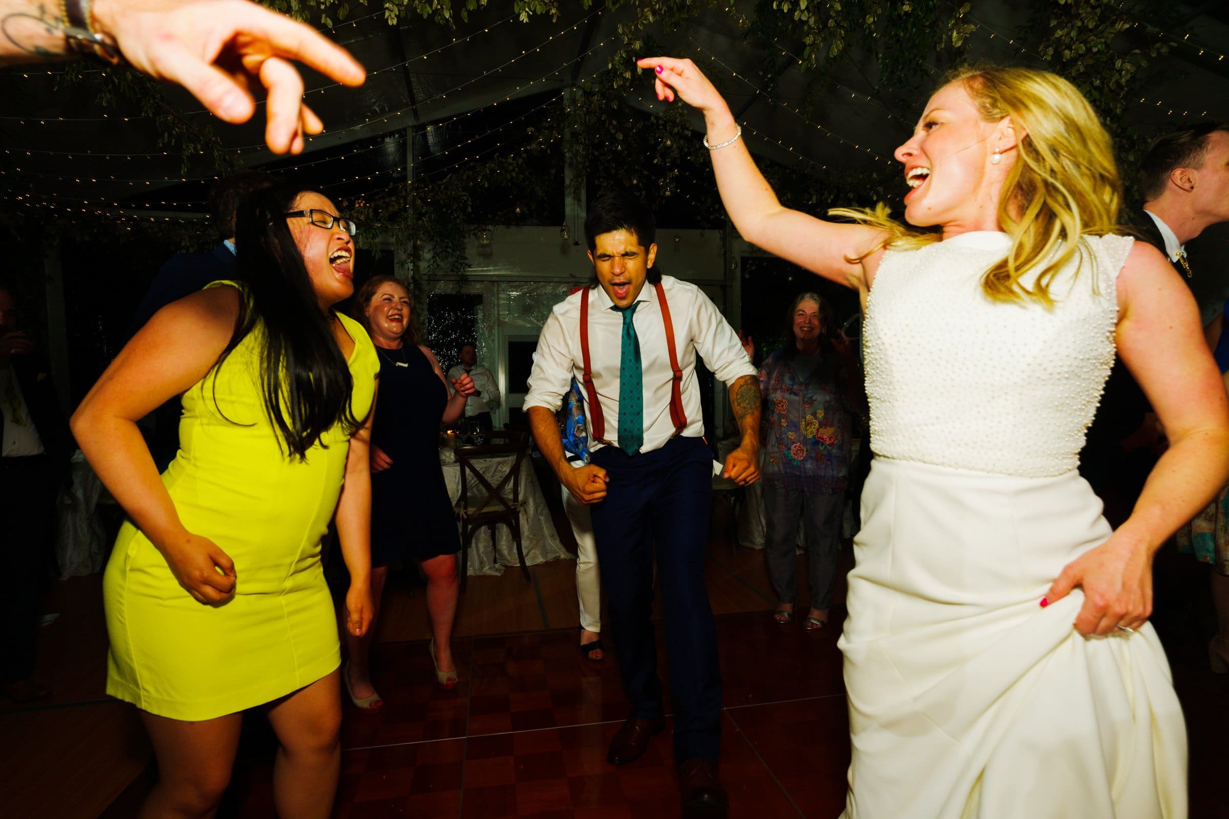 duke university wedding photography - sarah & case A joyful group of people dance energetically at a party, captured with the artistry of Duke University wedding photography. A woman in a white dress and a man in a shirt and tie stand out among others in vibrant outfits, all under festive lighting and lush greenery overhead.