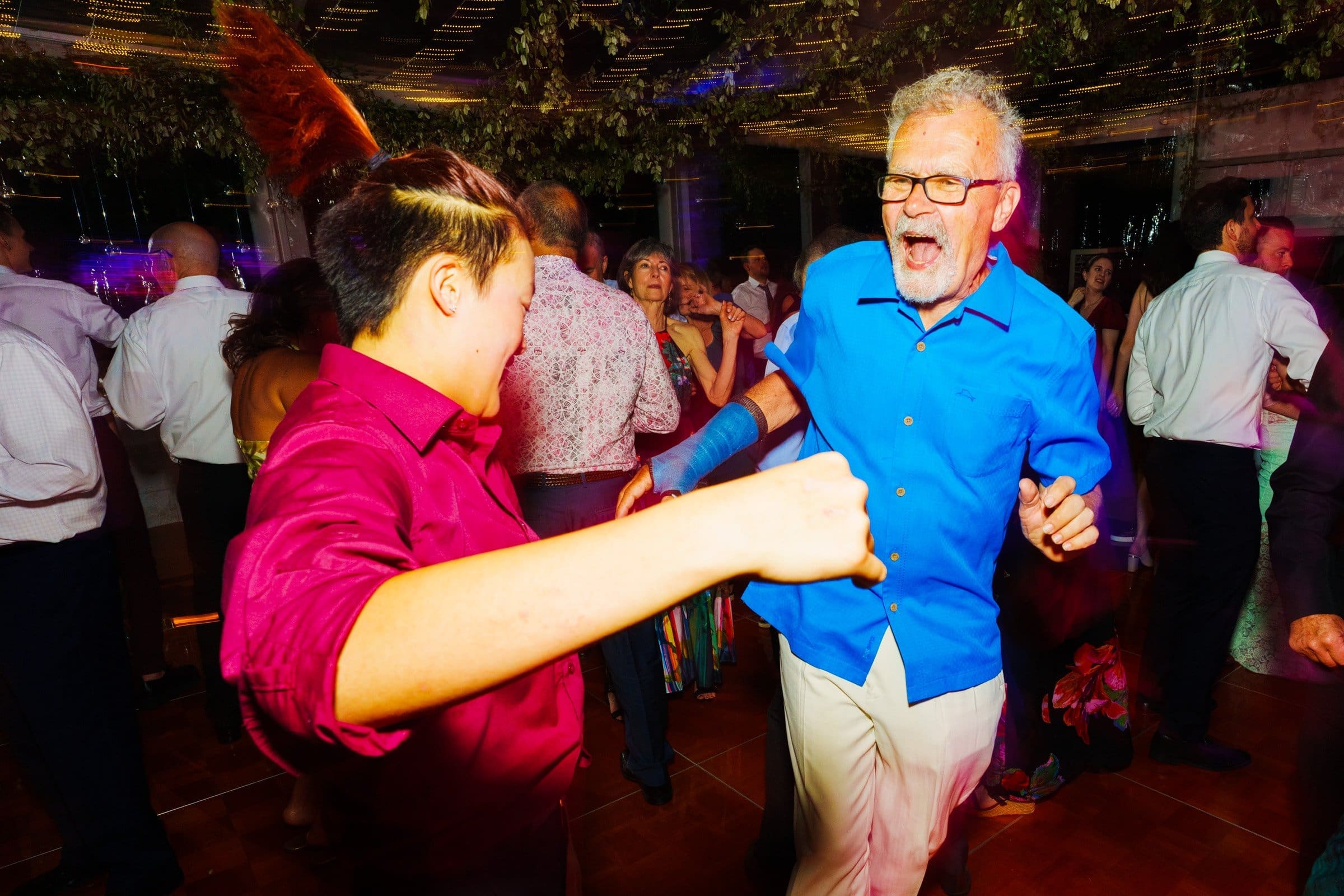 duke university wedding photography - sarah & case Two people dancing energetically at a lively party look like a scene straight out of a Duke University event. The person on the left sports a red shirt, while the one on the right dazzles in bright blue. Other guests mingle in the background beneath leafy decorations and colorful lighting, reminiscent of wedding photography.