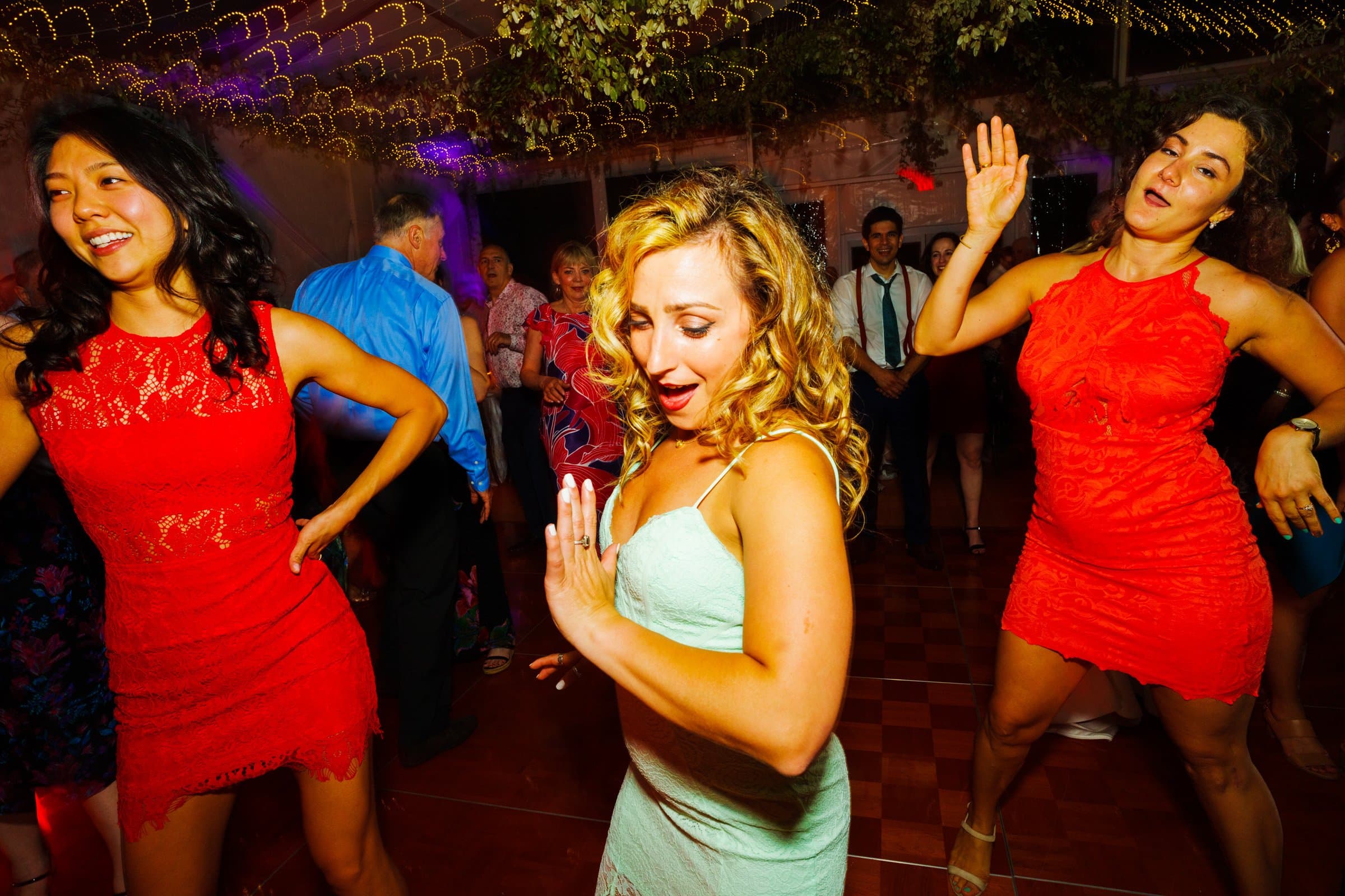 duke university wedding photography - sarah & case Three women are dancing energetically at a party, wearing brightly colored dresses. The vibrant scene feels reminiscent of Duke University's spirited events, with dim festive lighting and string lights overhead enhancing the lively atmosphere captured beautifully in wedding photography.