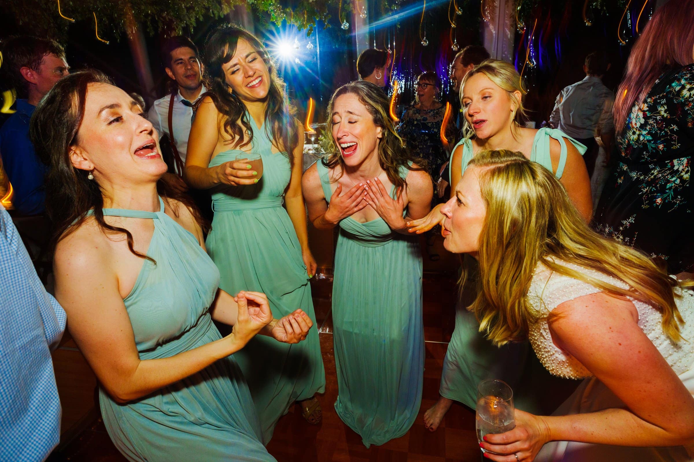 duke university wedding photography - sarah & case Five women in teal dresses joyfully sing and dance together on the vibrant dance floor at a lively event. Captured with Duke University precision, this wedding photography scene showcases a festive atmosphere surrounded by people and colorful lights.