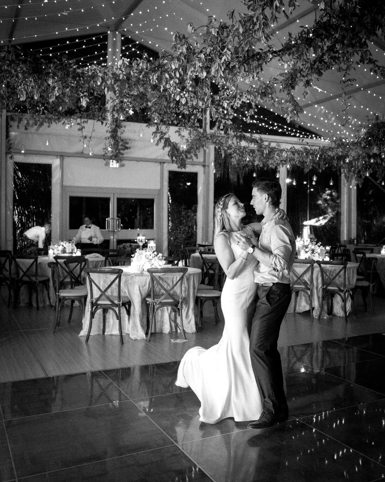 duke university wedding photography - sarah & case A couple dances gracefully in an elegantly decorated venue with hanging greenery and twinkling lights. The woman, in a long dress, and the man, in shirt and trousers, move effortlessly as if captured by Duke University wedding photography. Tables draped in tablecloths dot the background.