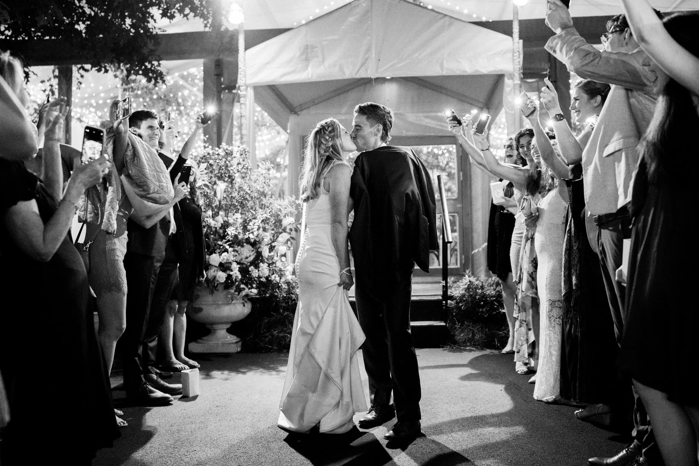 duke university wedding photography - sarah & case A newlywed couple walks hand in hand through a cheering crowd at night, captured elegantly by Duke University wedding photography. The bride wears a long dress, and the groom is in a suit. Guests hold sparklers, creating a festive atmosphere as string lights illuminate the scene.
