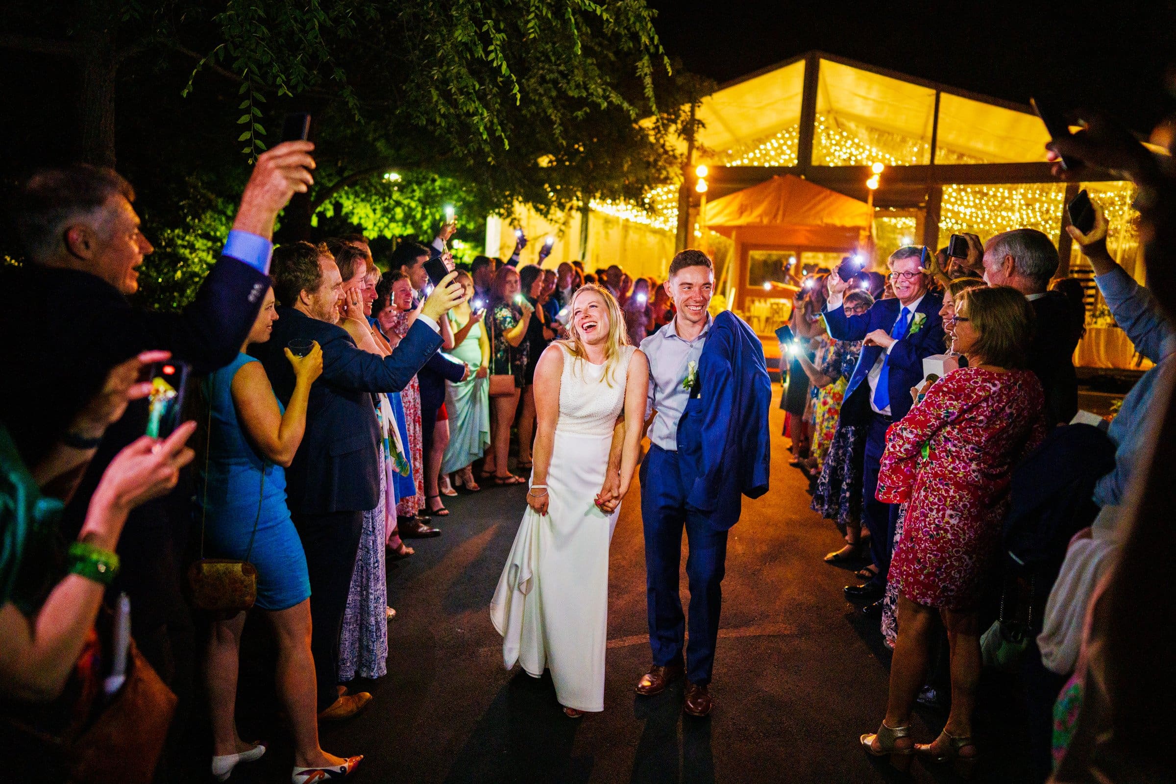 duke university wedding photography - sarah & case A joyful couple, dressed in wedding attire, walks hand-in-hand down a pathway lined with smiling guests. The scene is illuminated by warm glowing lights strung above, capturing a festive and celebratory atmosphere at night—an unforgettable moment of Duke University wedding photography.